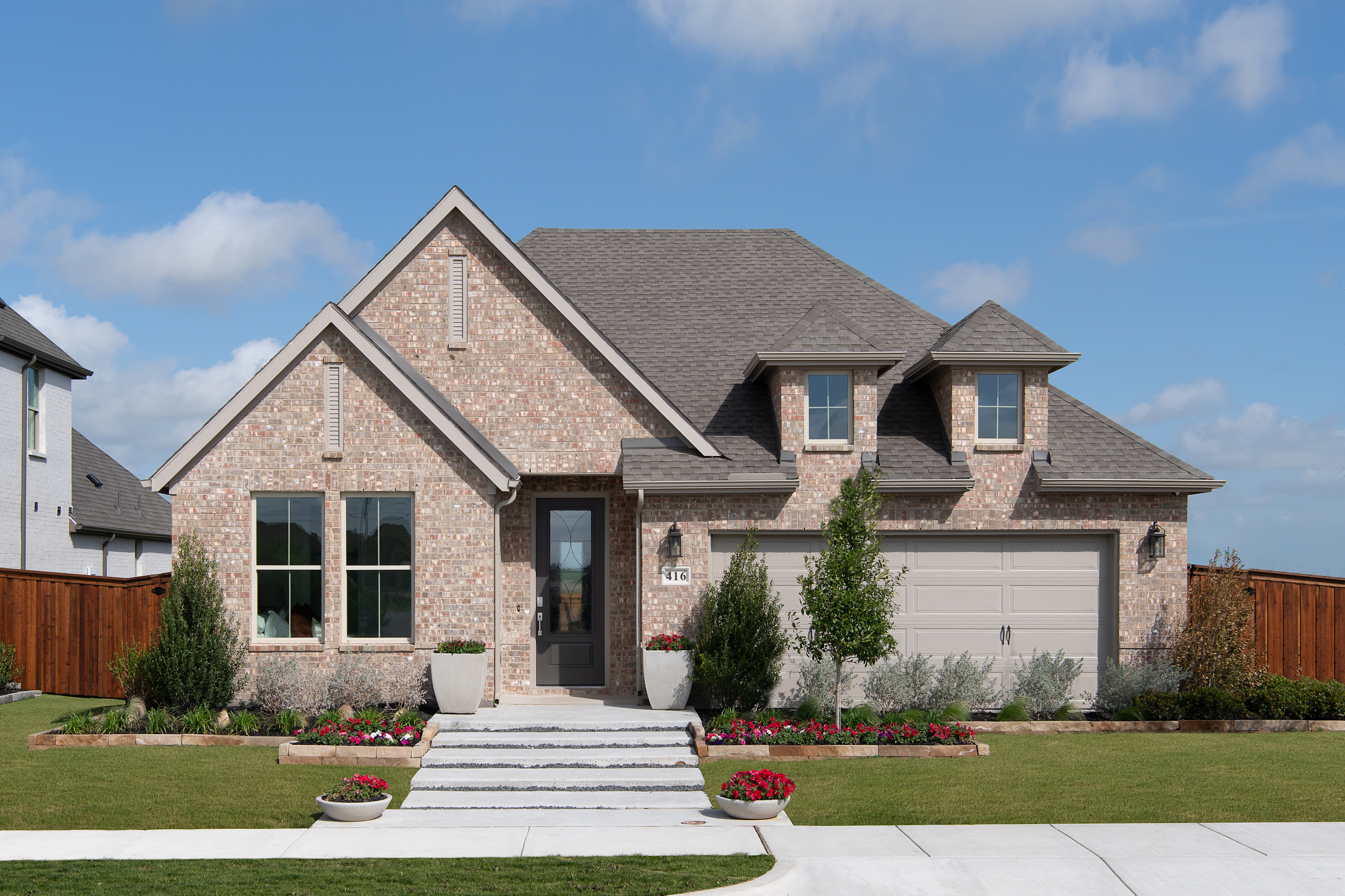 A two-story brick house with a gabled roof, surrounded by a well-manicured lawn and landscaped garden, set against a blue sky with scattered clouds.