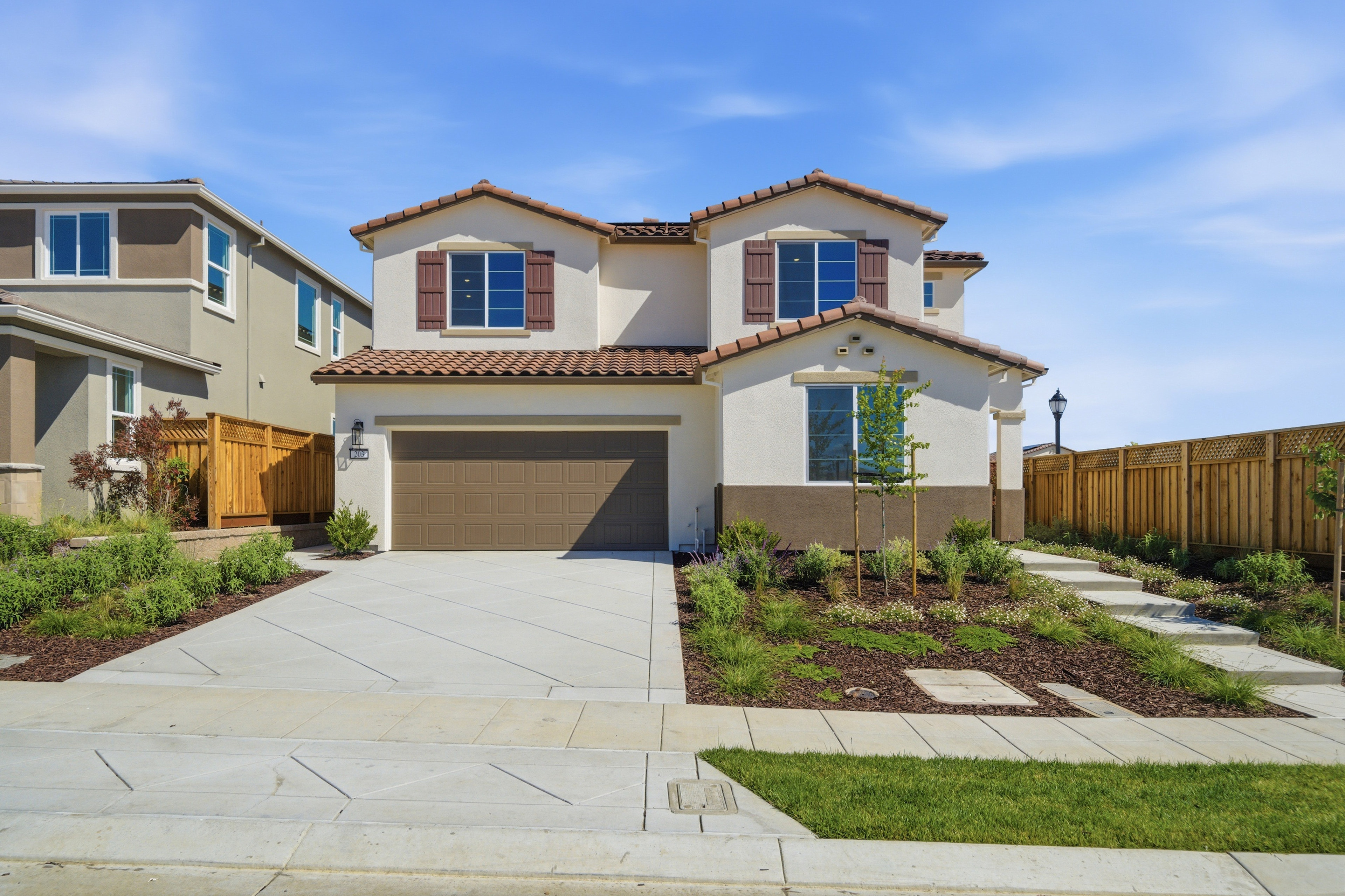 A two-story Mediterranean-style house with a tiled roof, surrounded by a well-manicured lawn, paved walkway, and a wooden fence in the background.