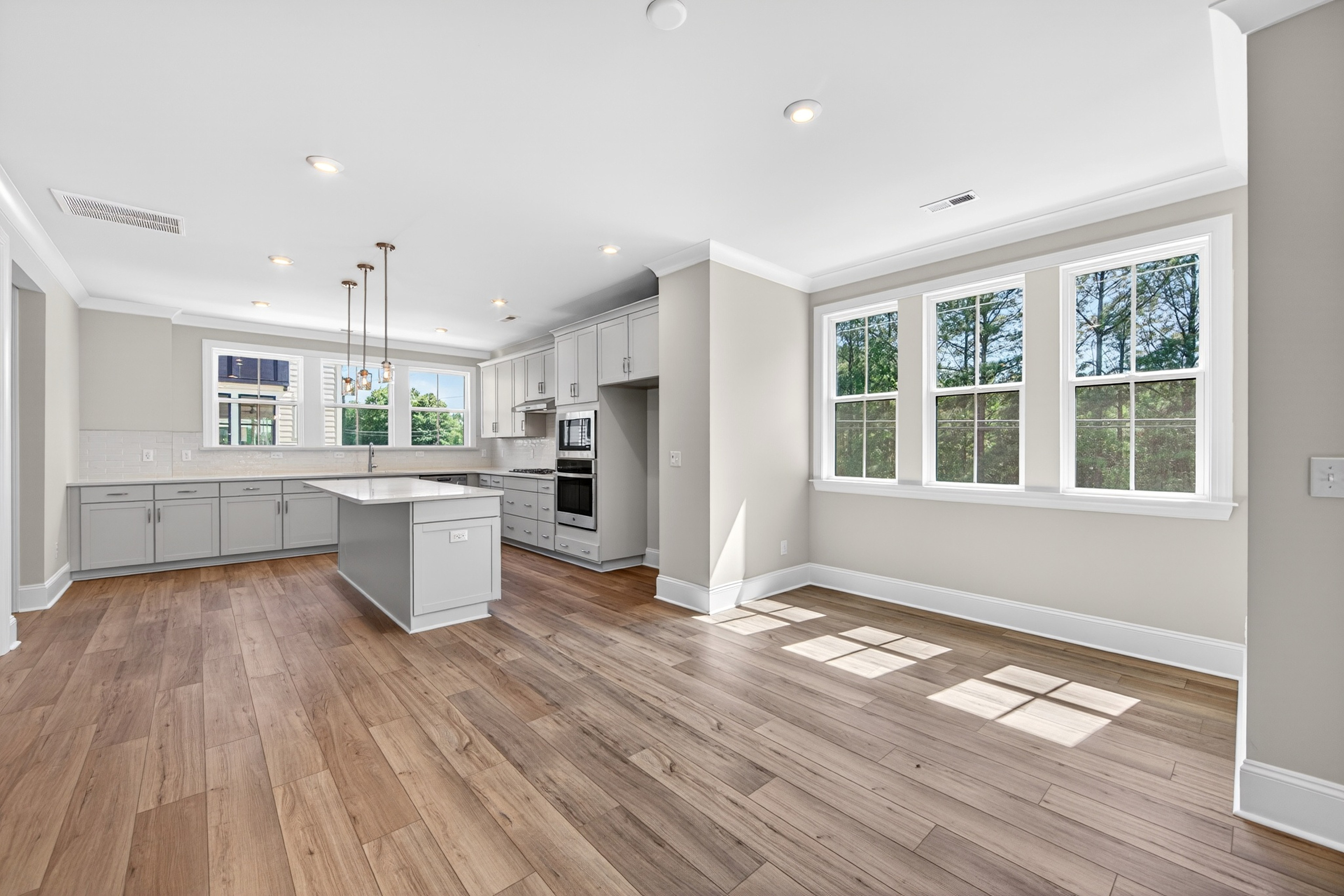 A modern, open-concept kitchen with white cabinets, stainless steel appliances, and large windows overlooking a wooded outdoor area.