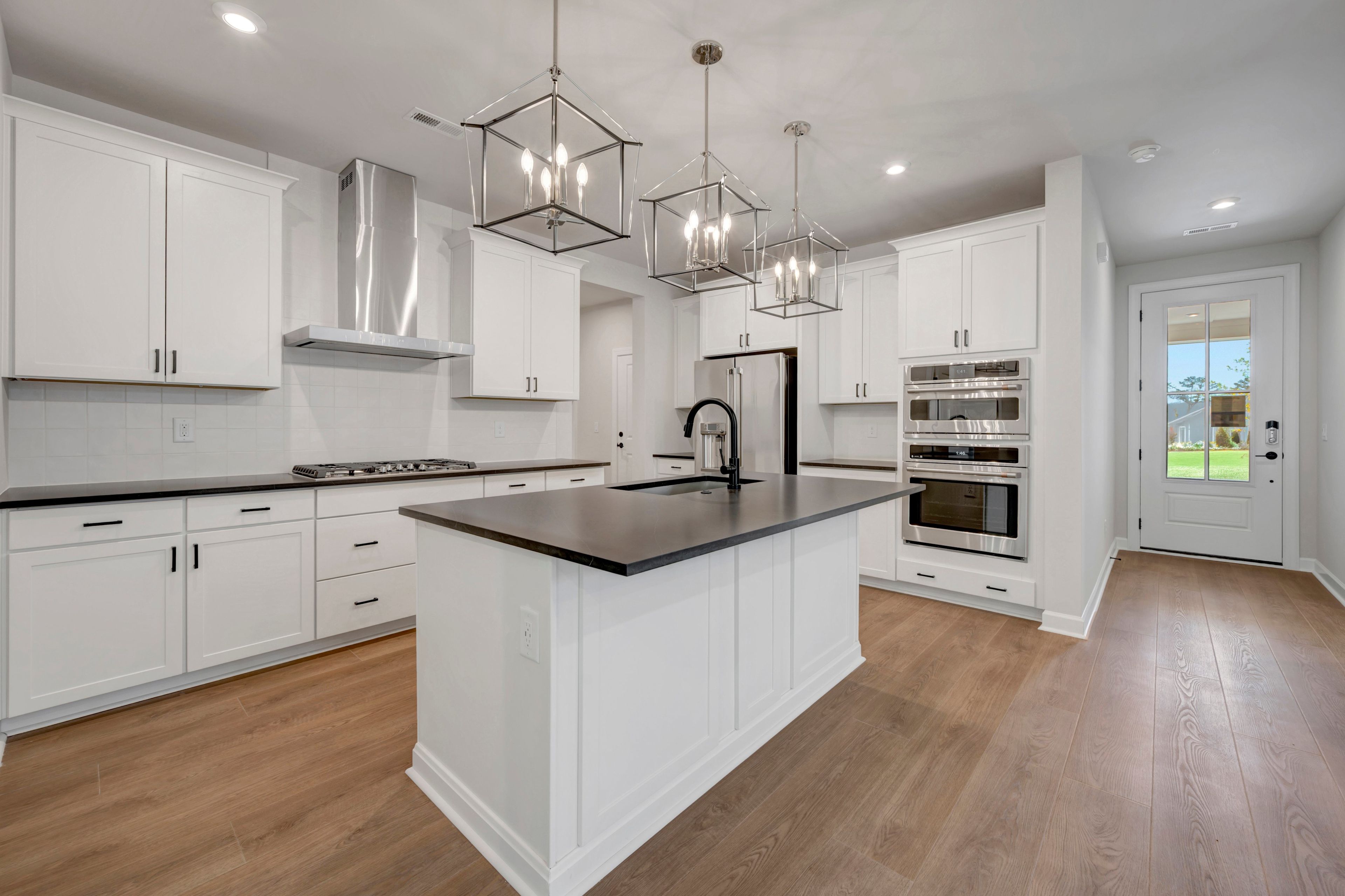 A modern, well-lit kitchen with white cabinets, a dark countertop, and pendant lighting fixtures, leading to a hallway with a glass door.