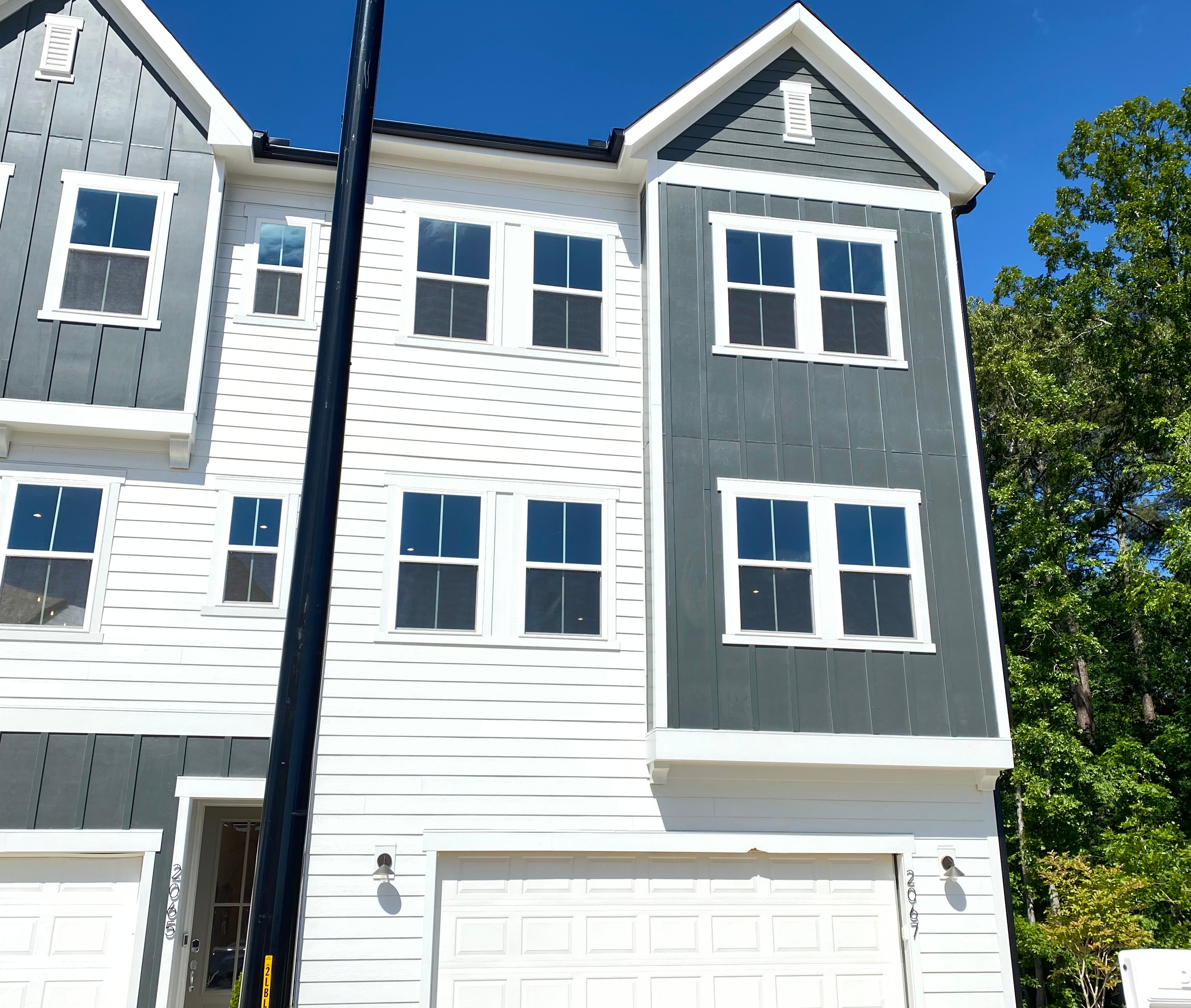 A two-story townhouse with white siding and gray trim, surrounded by trees and a clear blue sky.