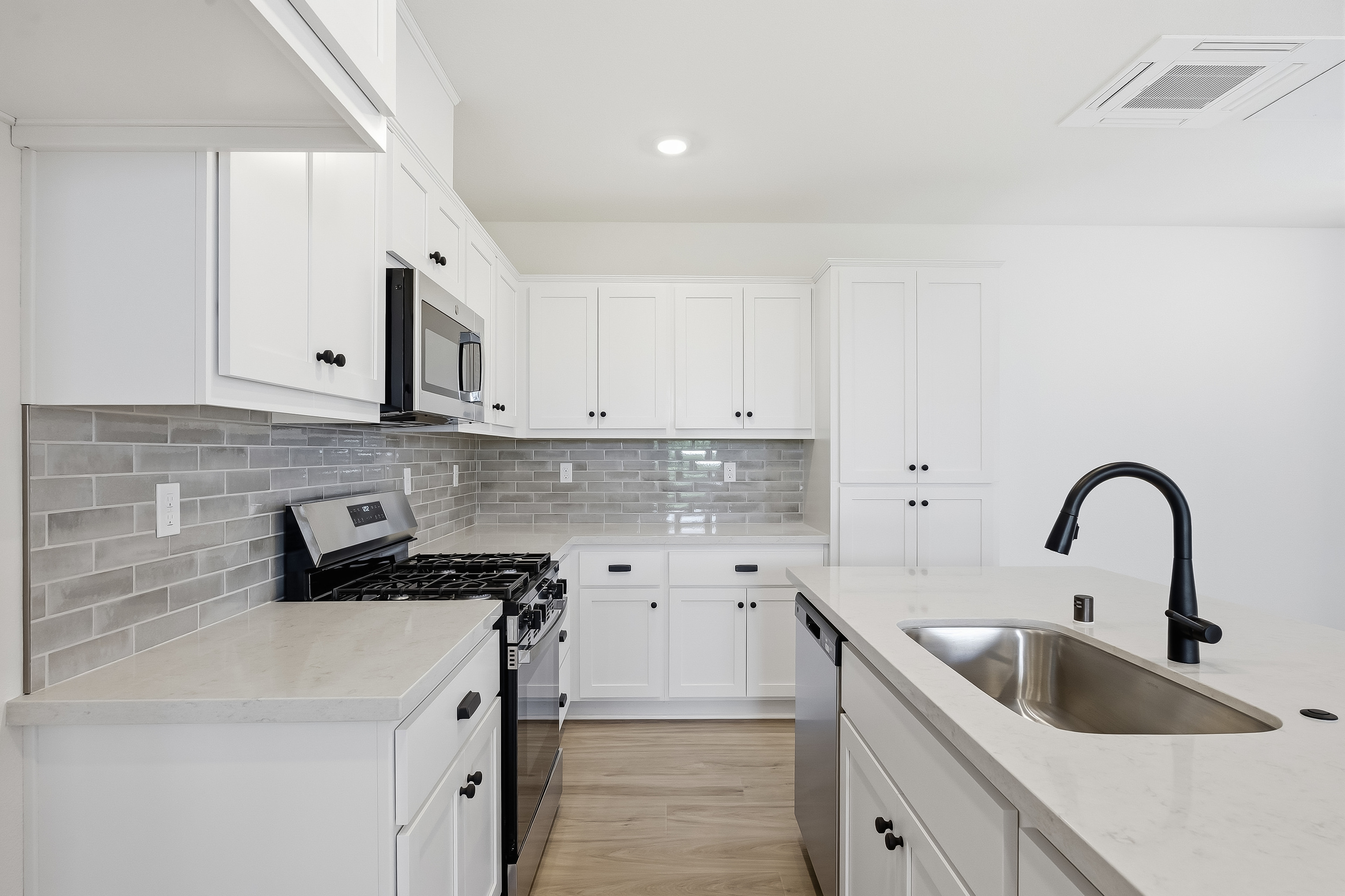 A modern white kitchen with a gray subway tile backsplash and a black faucet over a stainless steel sink in the foreground, and white cabinets and appliances in the background.