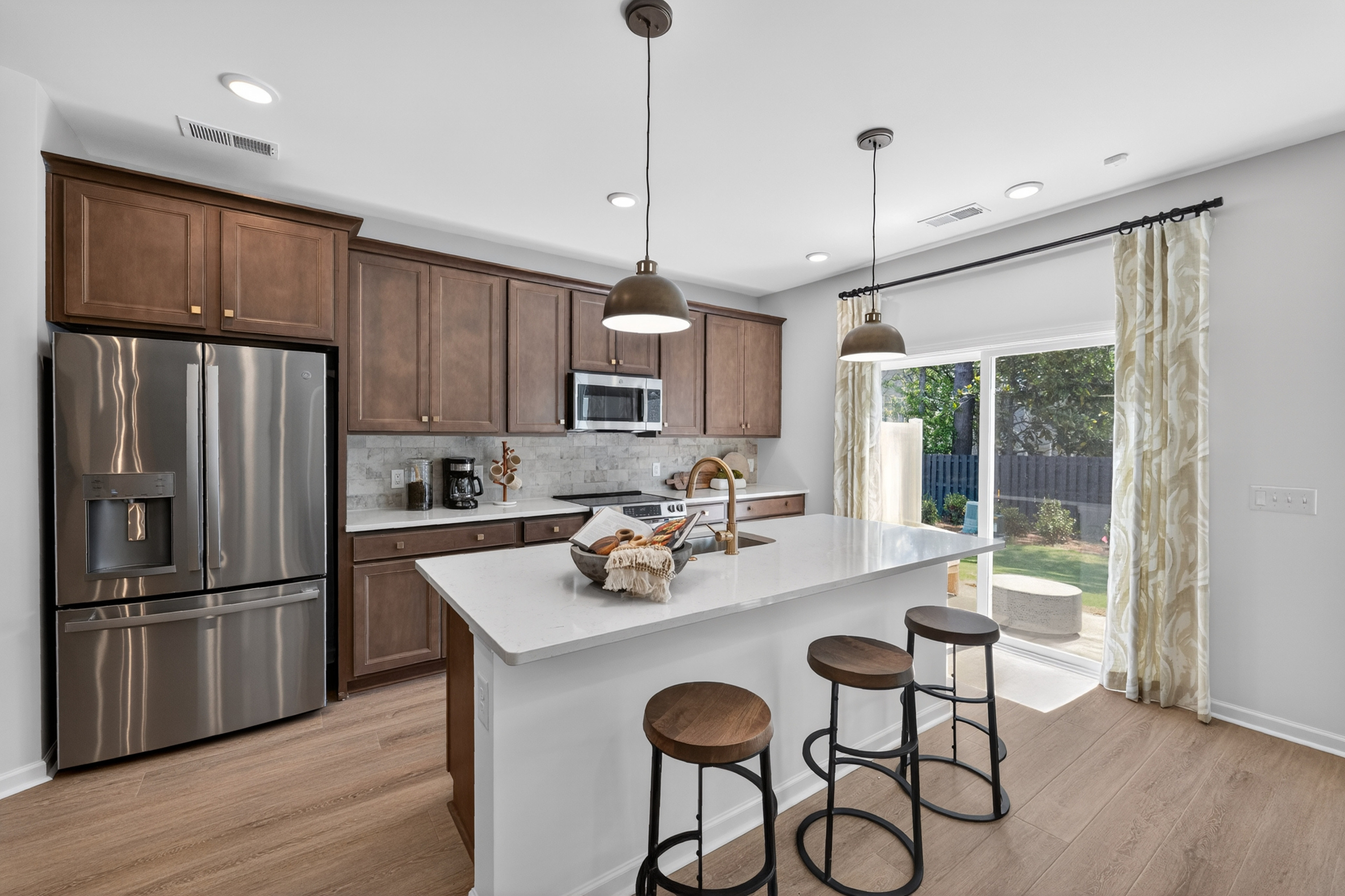 A modern kitchen with a stainless steel refrigerator and island with bar stools in the foreground, and a sliding glass door leading to a backyard in the background.