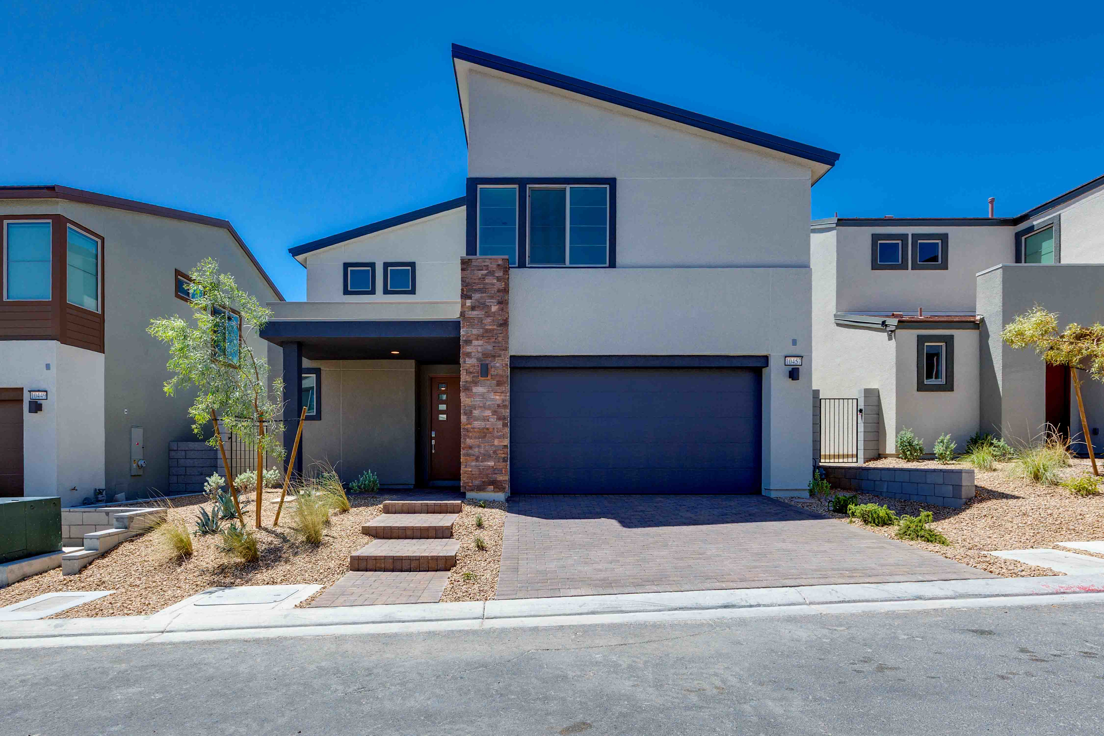 A modern, two-story house with a dark garage door and a brick-accented entrance stands on a paved driveway, with other contemporary homes and a clear blue sky in the background.