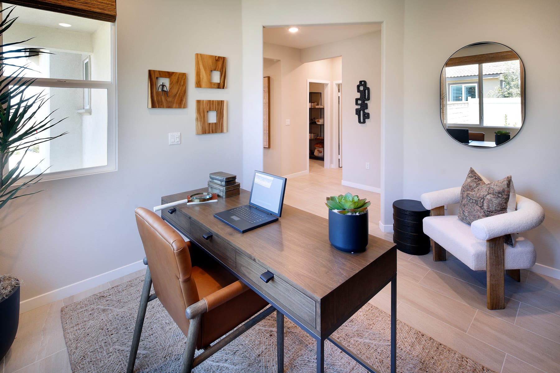A cozy and well-designed home office space with a wooden desk, laptop, and decorative elements like wall art and a potted plant, set against a neutral-toned background with a hallway visible in the distance.