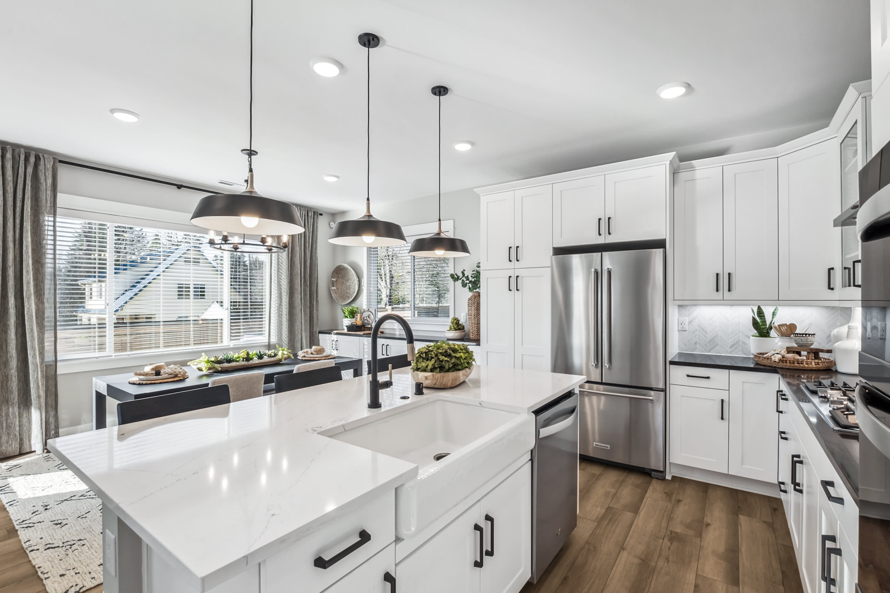 A modern white kitchen with a large island in the foreground and a dining area with a window overlooking houses in the background.