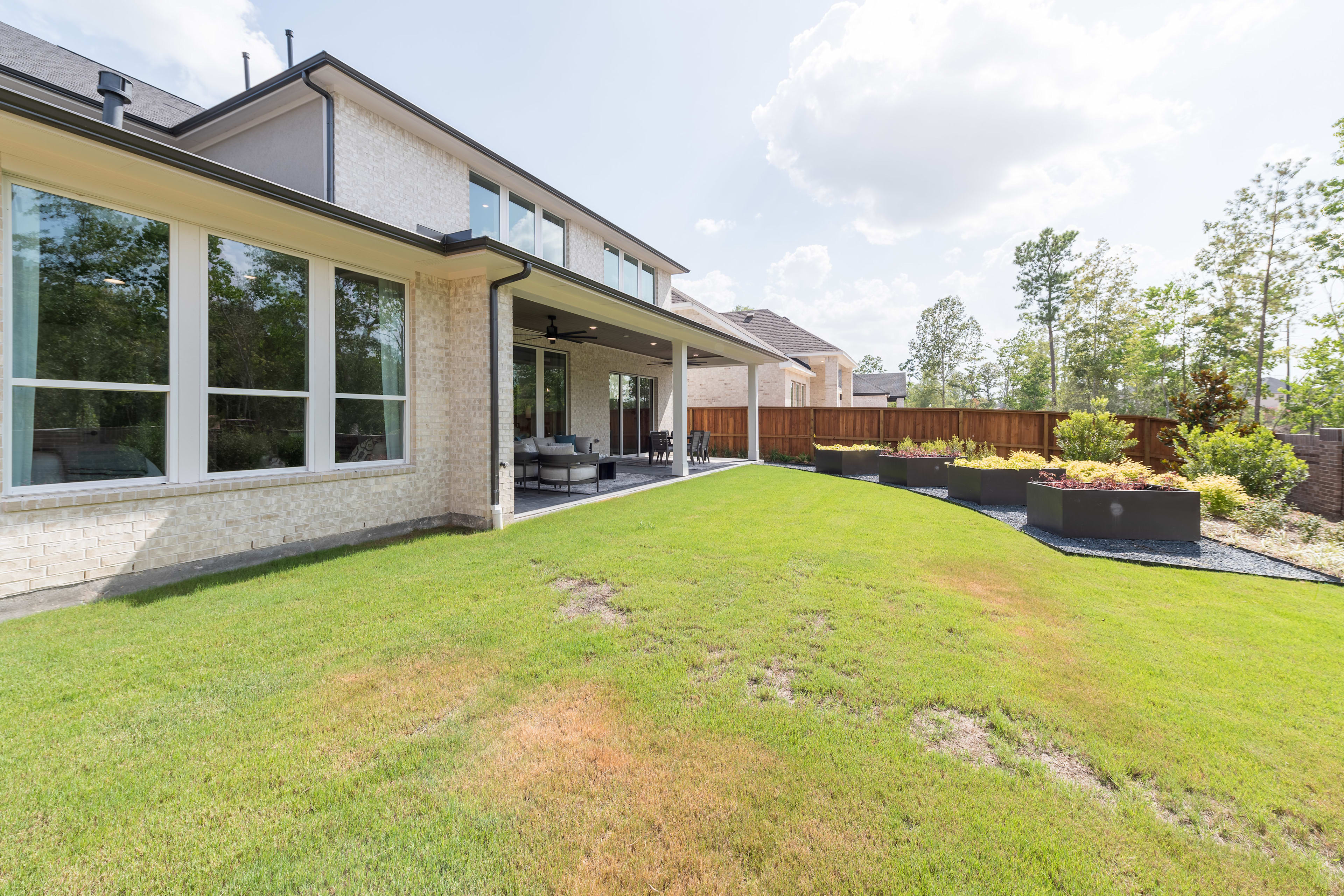 A well-manicured lawn leads to a modern, two-story house with large windows and a covered patio, surrounded by lush greenery and trees.