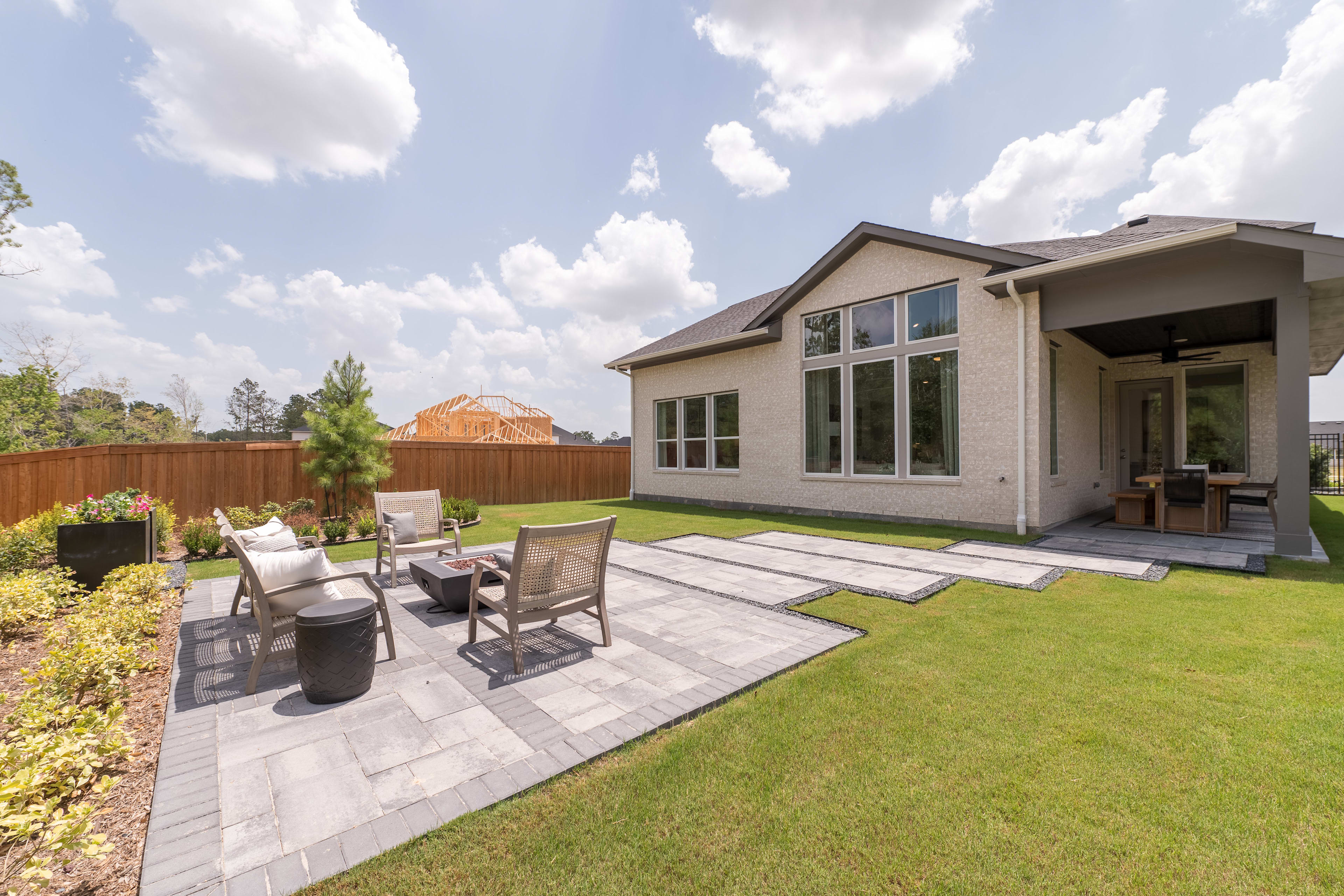 A modern, two-story house with a large patio and well-manicured lawn in the foreground, surrounded by lush greenery and a cloudy sky in the background.