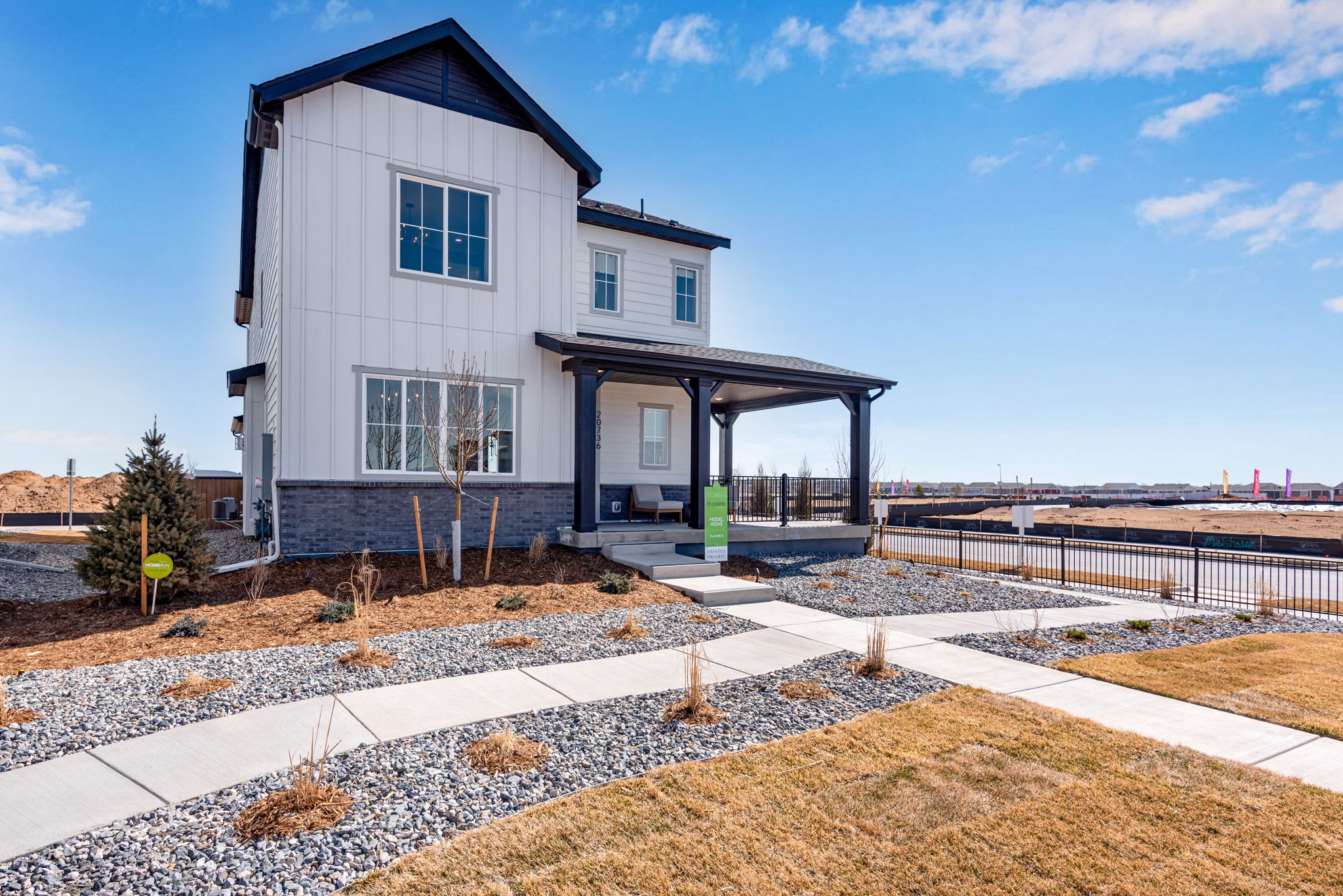 A modern, two-story white and gray house with a covered porch sits on a landscaped lot with a gravel path leading to the front door, surrounded by a clear blue sky with scattered clouds.