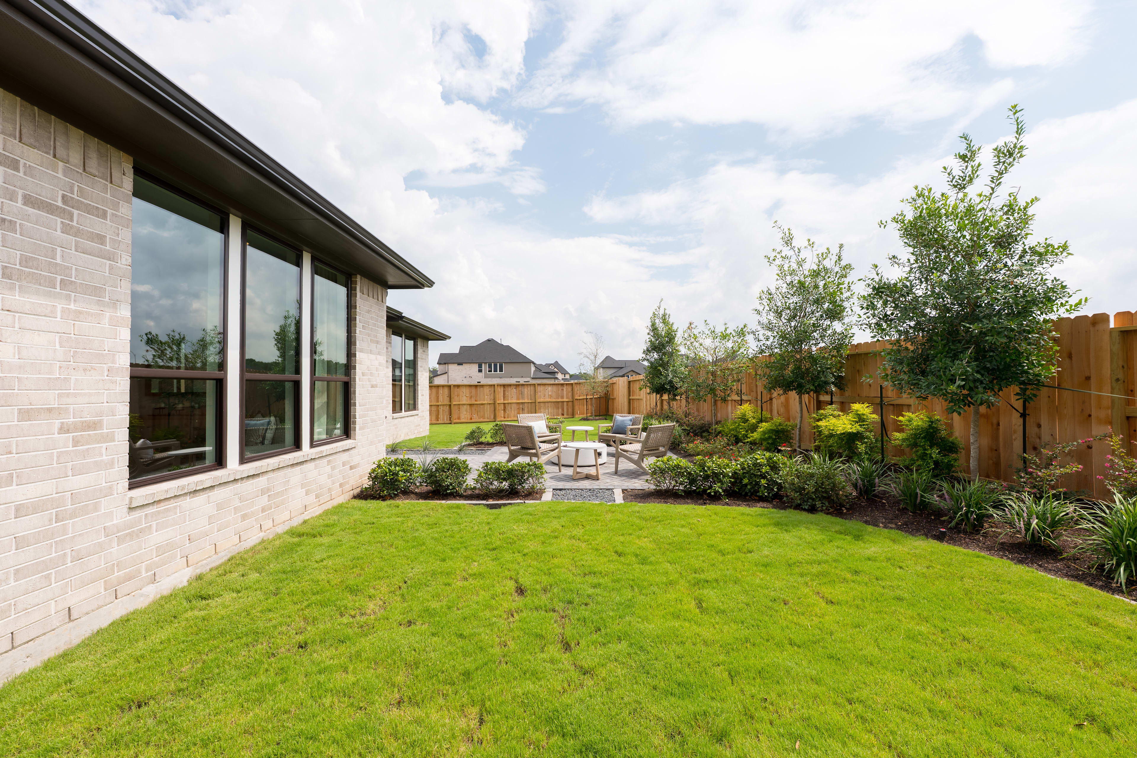 A well-manicured backyard with a lush green lawn, surrounded by a wooden fence and featuring a patio area with outdoor furniture, set against a backdrop of a modern brick building with large windows.
