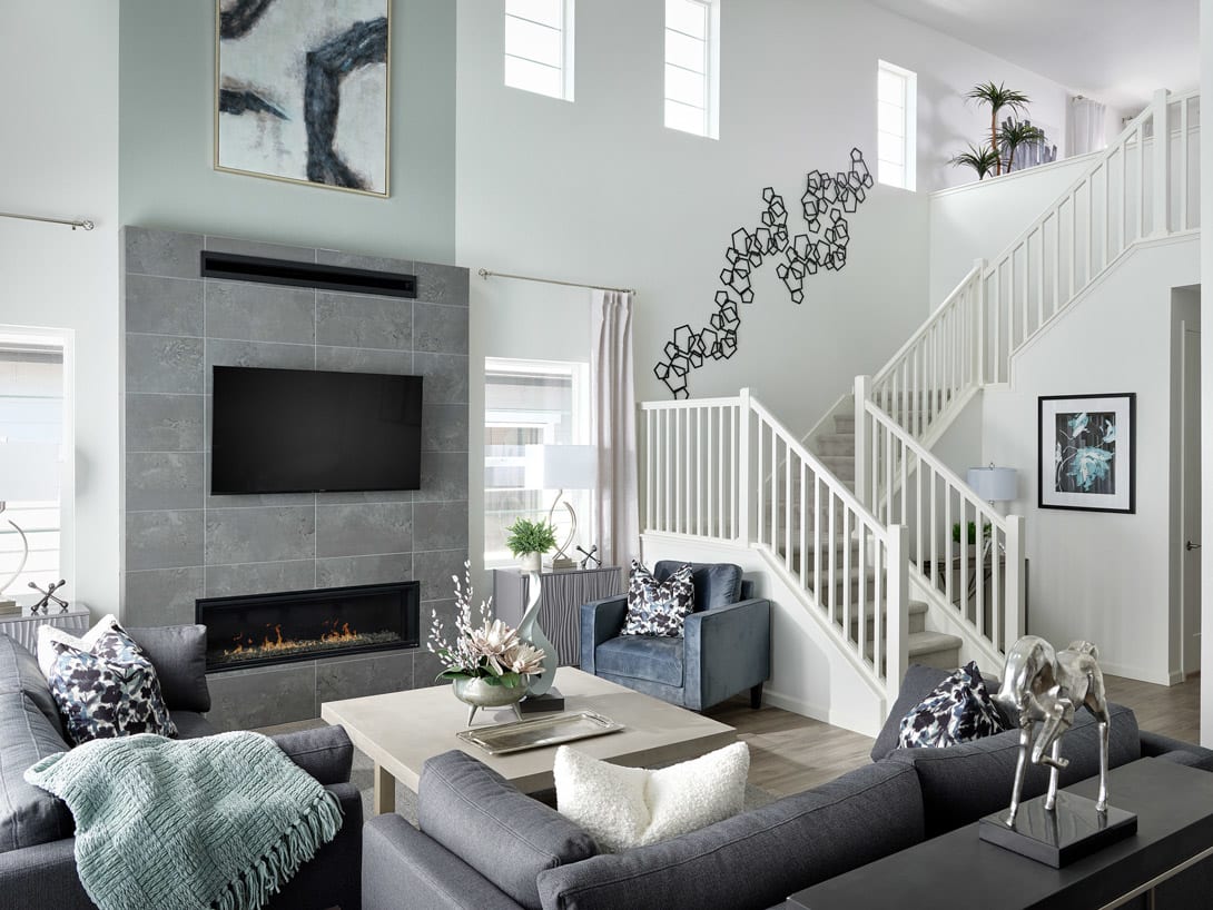 A modern and minimalist living room with a fireplace, gray sofas, and a staircase leading to the upper level, surrounded by white walls and large windows allowing natural light to fill the space.