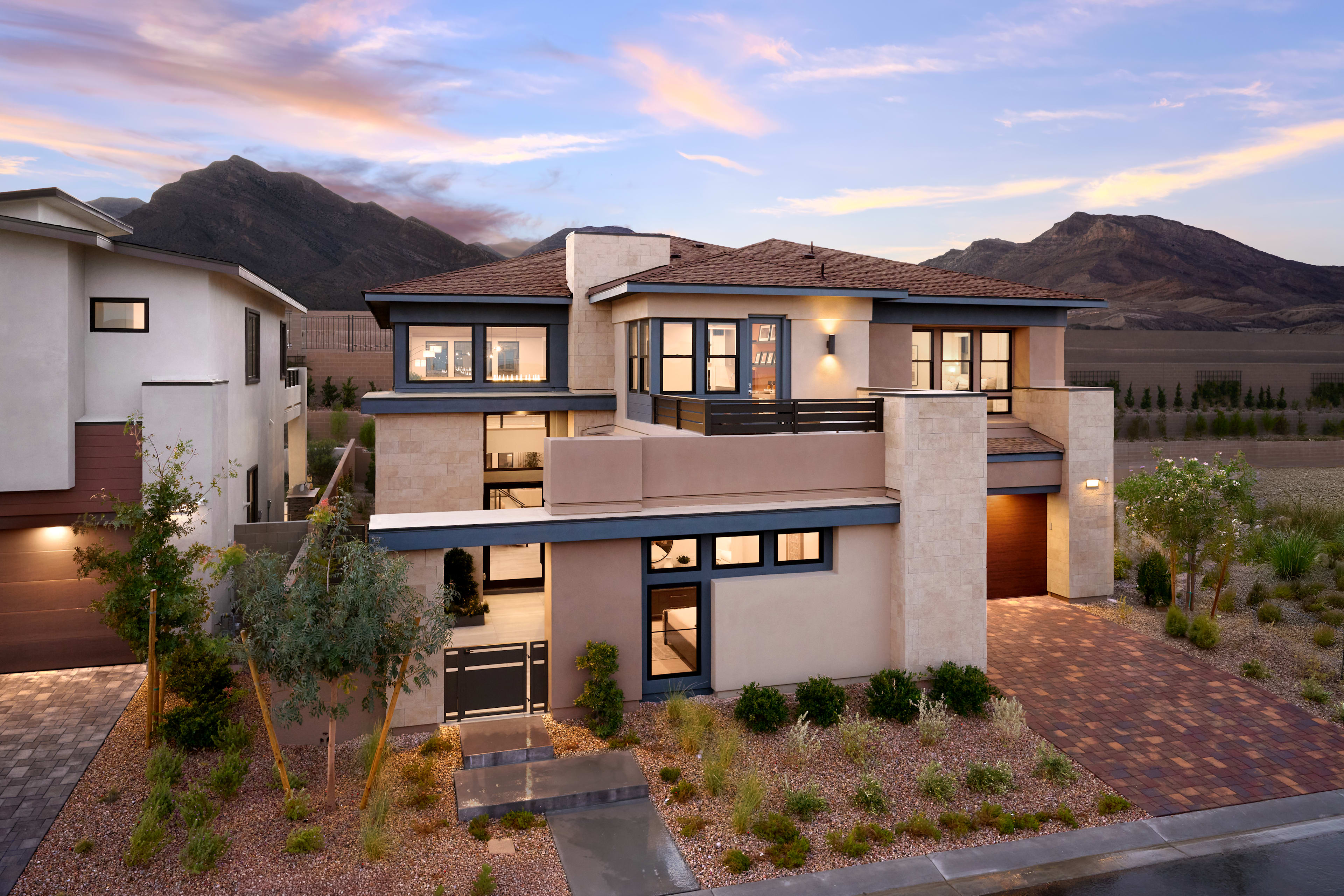 A modern, multi-story house nestled in a desert landscape, with mountains in the background and a paved driveway leading up to the entrance.