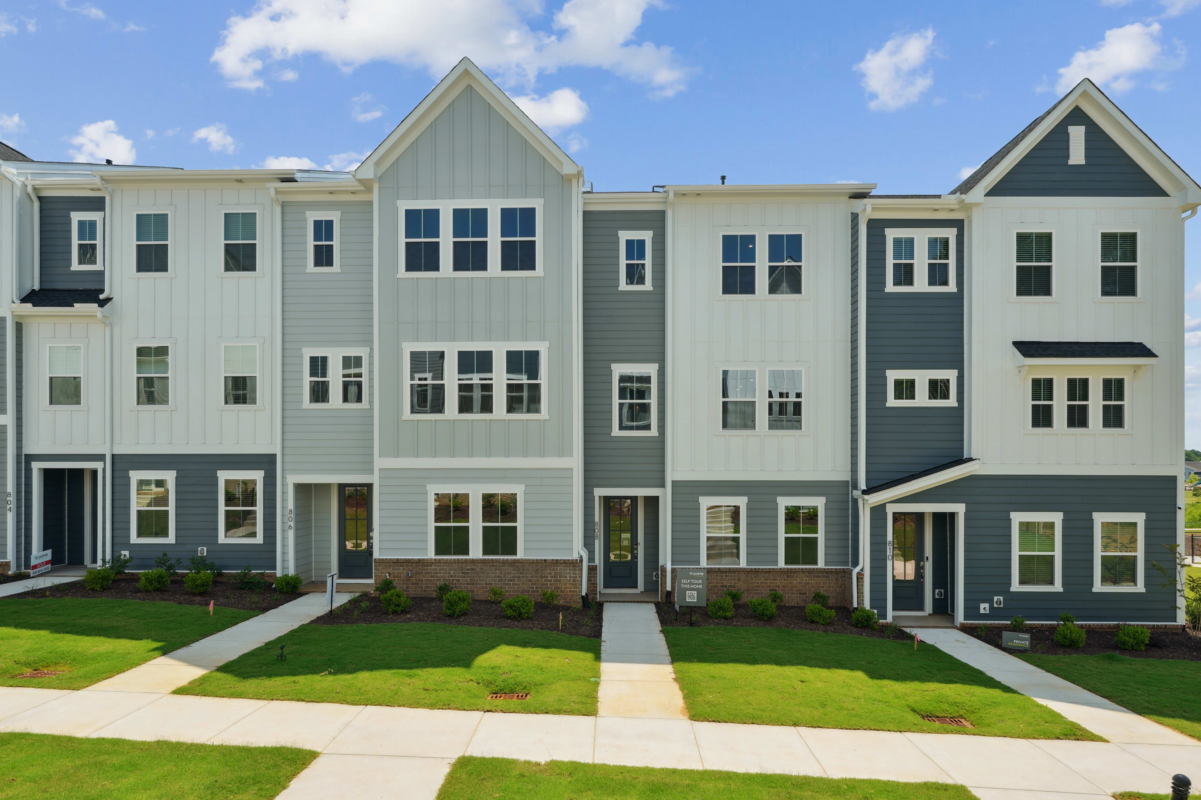 A row of modern, multi-story townhouses with gray siding, white trim, and gabled roofs, set against a backdrop of a blue sky with fluffy white clouds. The foreground features a well-manicured lawn with a paved walkway leading to the front entrances.