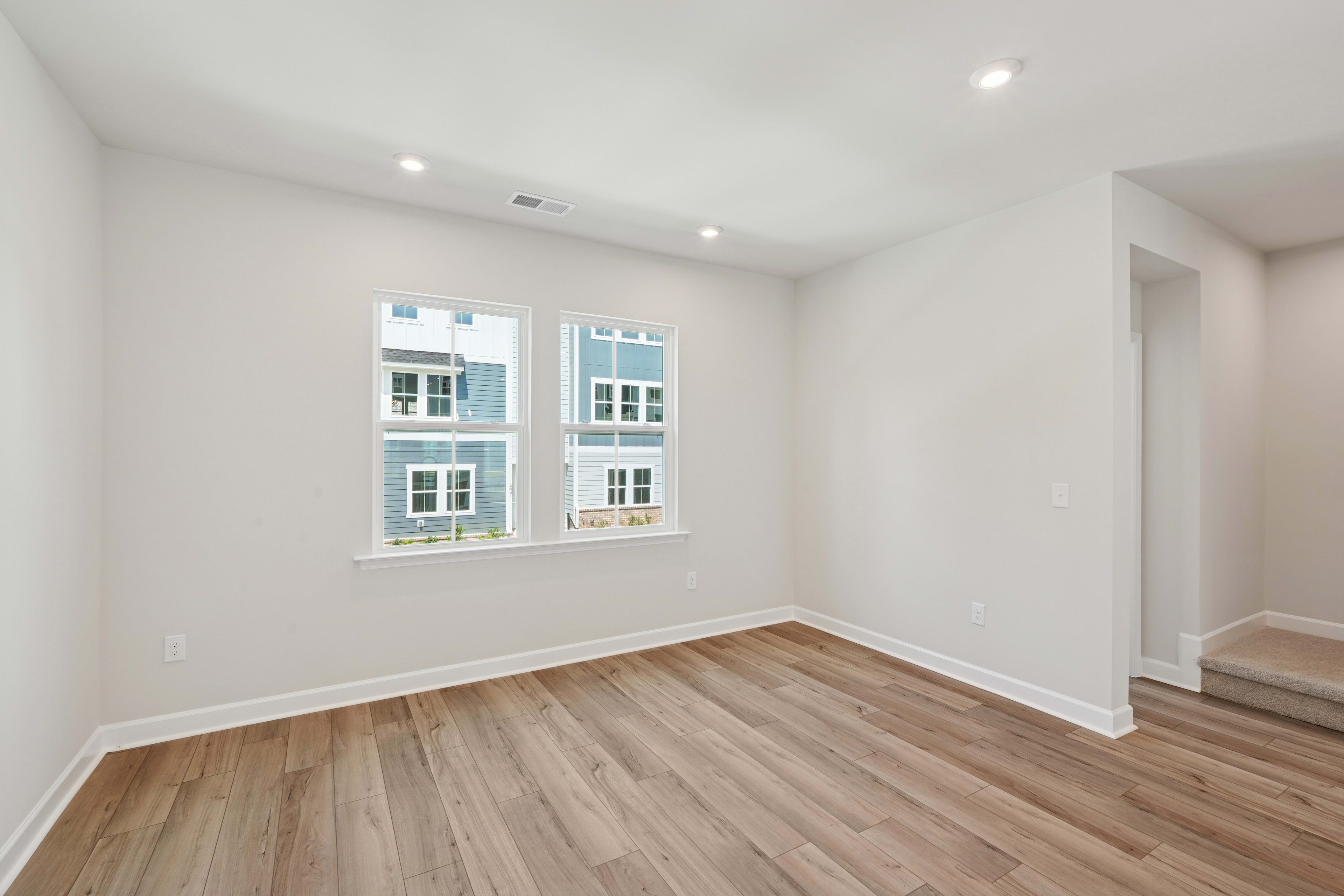 A spacious, well-lit room with white walls, wooden flooring, and large windows providing a view of the exterior.