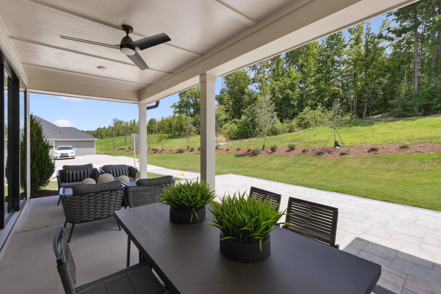 A covered patio with a ceiling fan, outdoor furniture, and a lush green lawn surrounded by trees in the background.