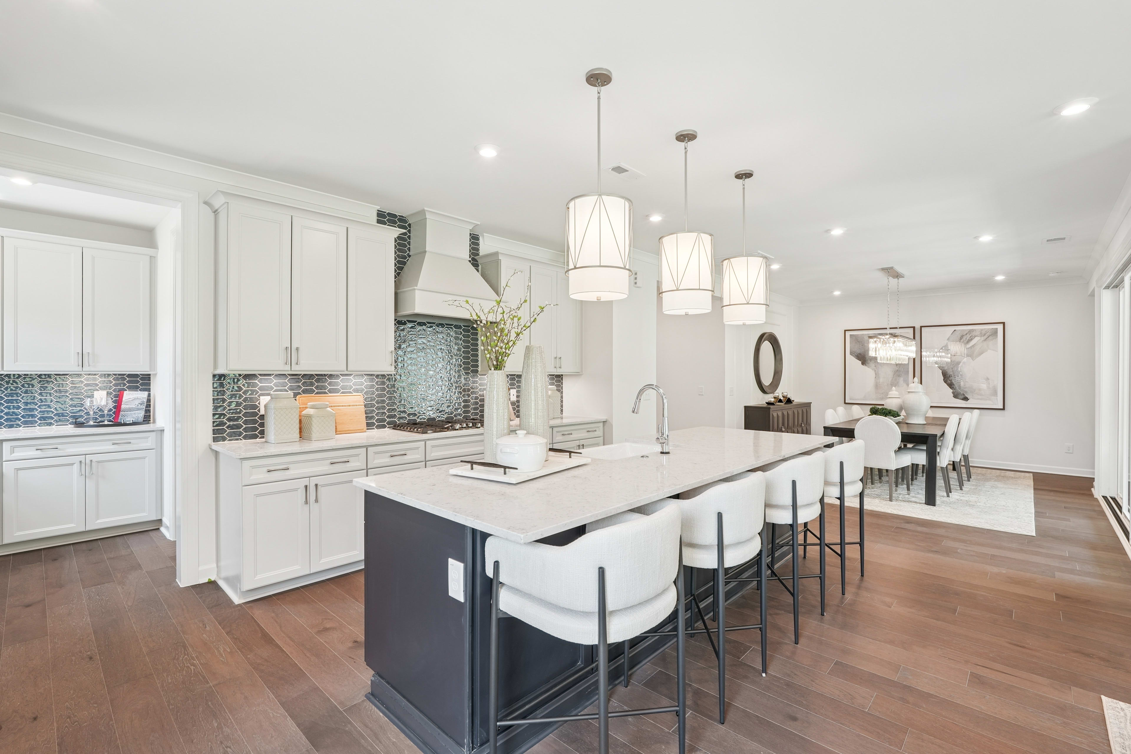 A modern, open-concept kitchen with white cabinets, a large island with bar stools, and pendant lighting fixtures, leading into a dining area with a wooden table and chairs.