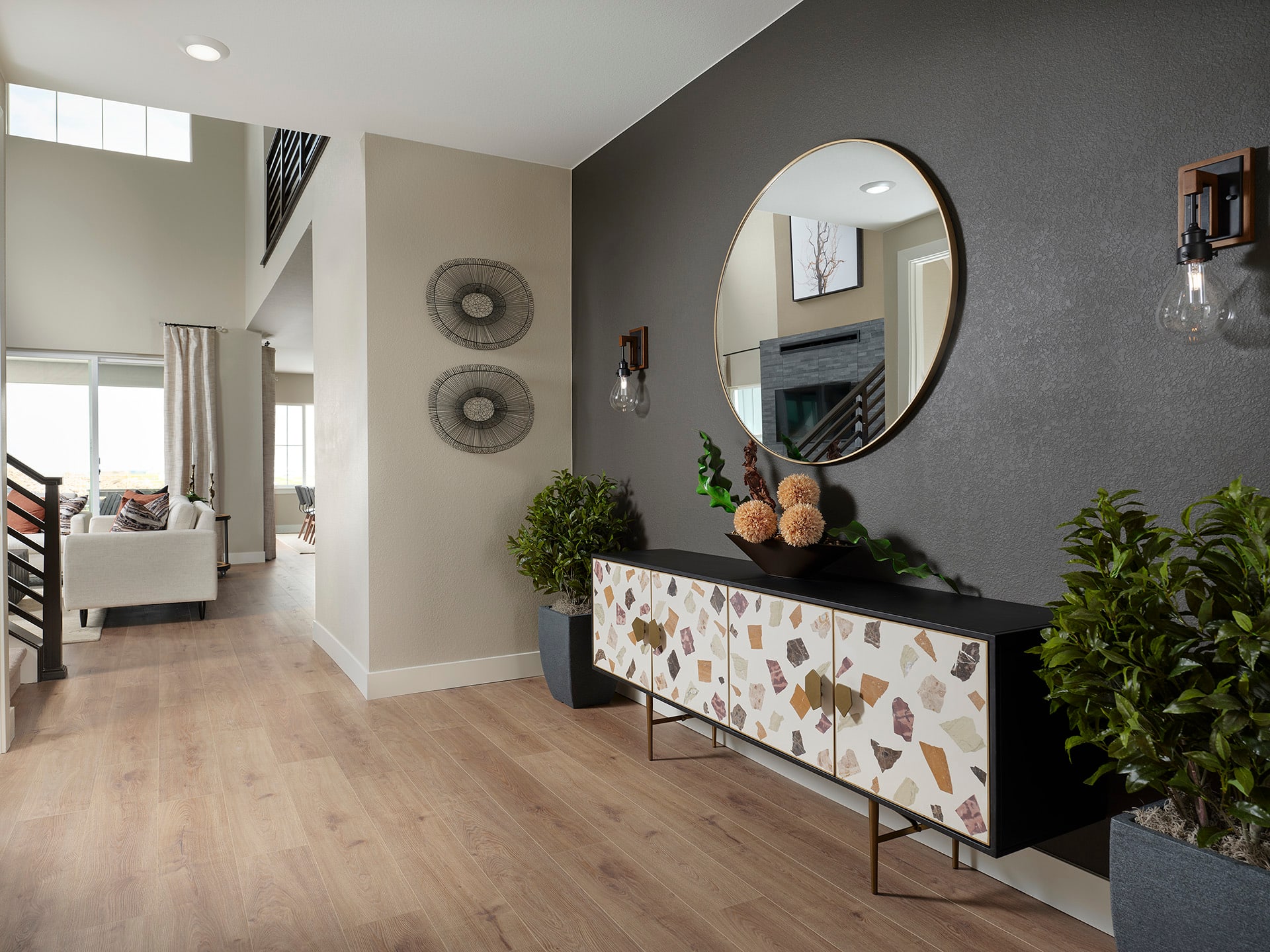 A modern and stylish entryway with a dark-colored accent wall, a round mirror, and a decorative console table with a mosaic pattern, surrounded by potted plants and other decor elements.