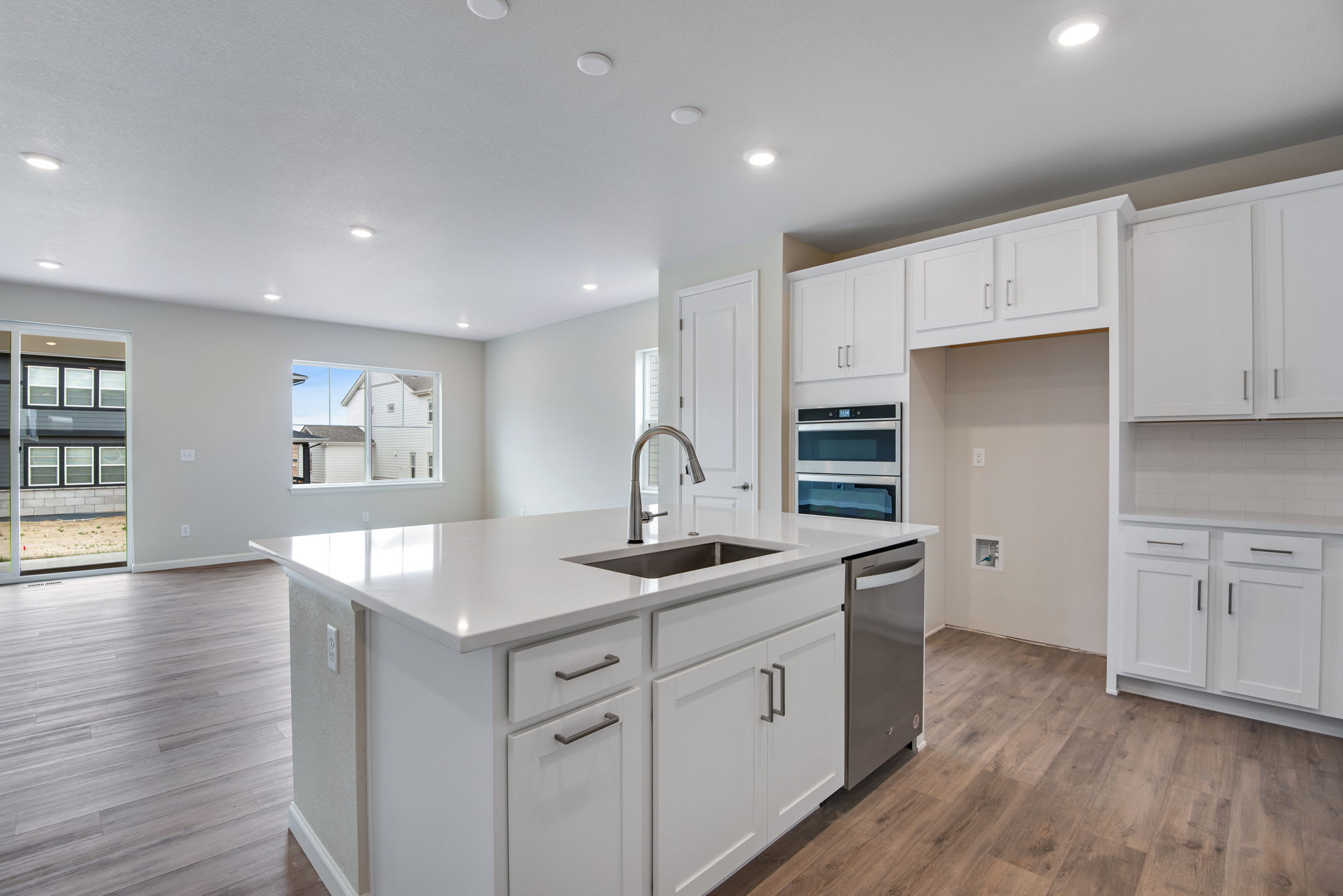 A modern, well-lit kitchen with white cabinets, a large island with a sink, and hardwood floors, set against a backdrop of windows and a bright, open living space.