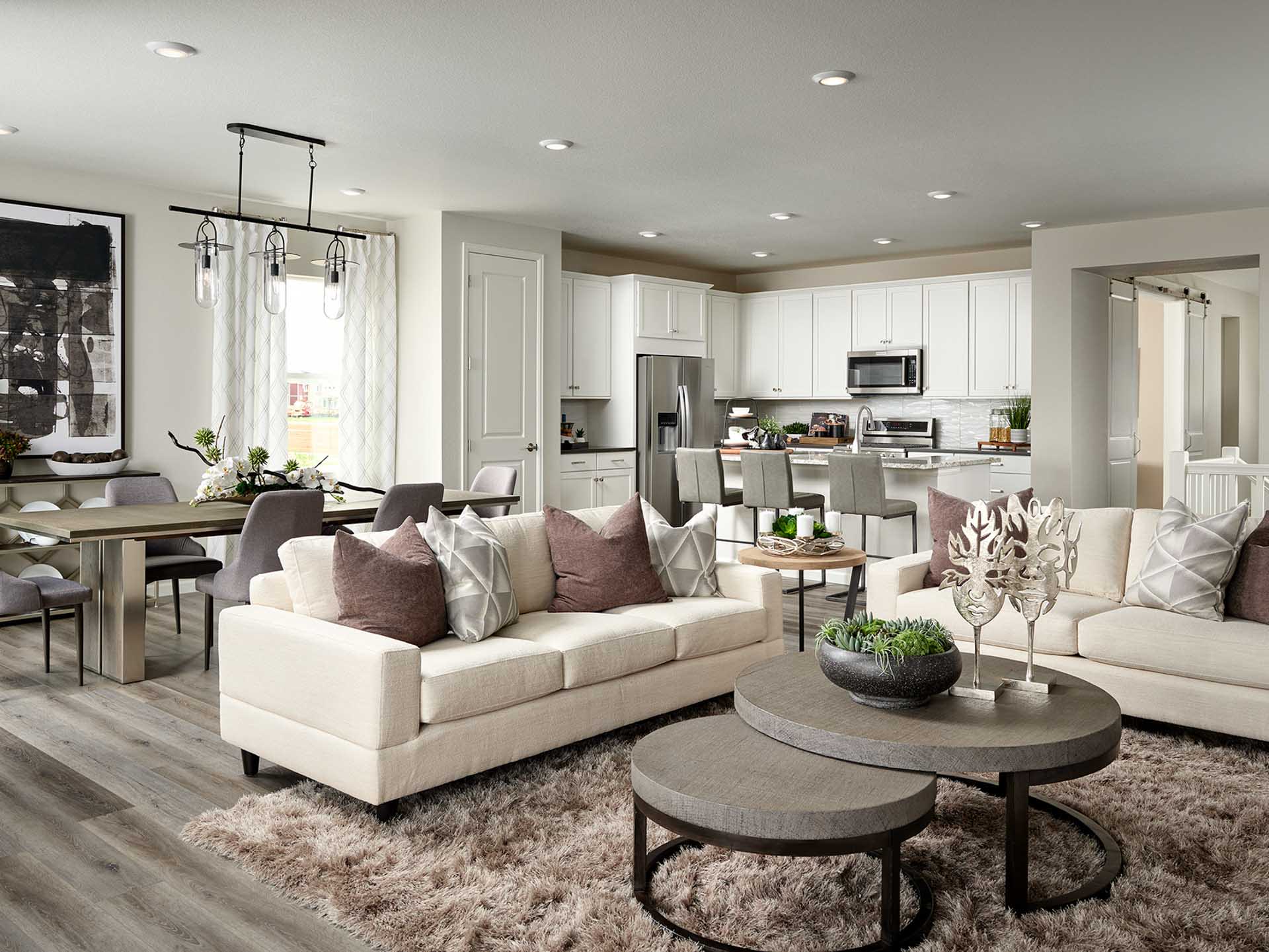 A cozy and modern living room with a plush white sofa, round coffee tables, and an open-concept kitchen visible in the background, all set against a neutral color palette and warm lighting.