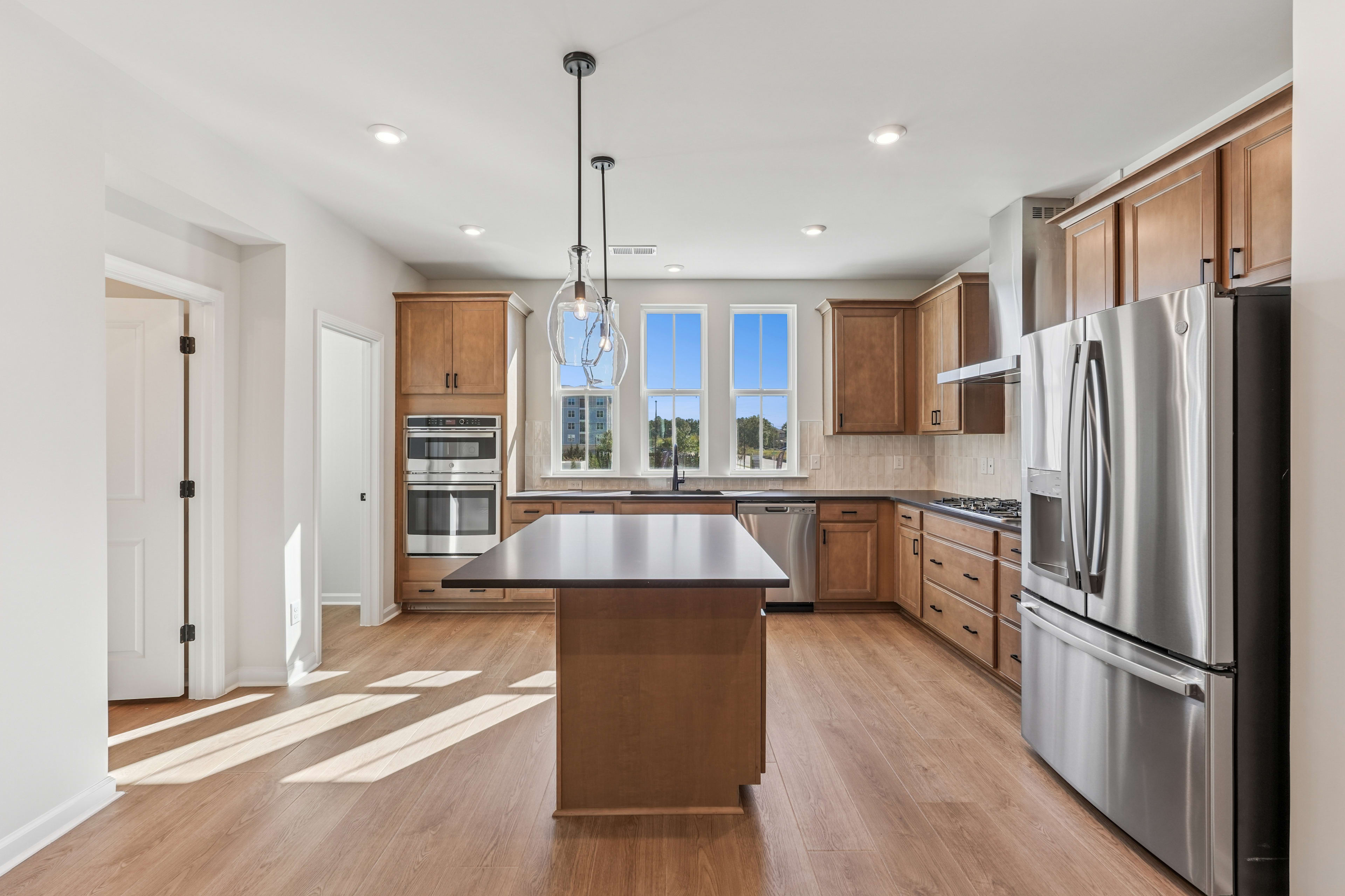 A modern, open-concept kitchen with wooden cabinets, stainless steel appliances, and a central island with a countertop, set against a backdrop of large windows providing natural light.