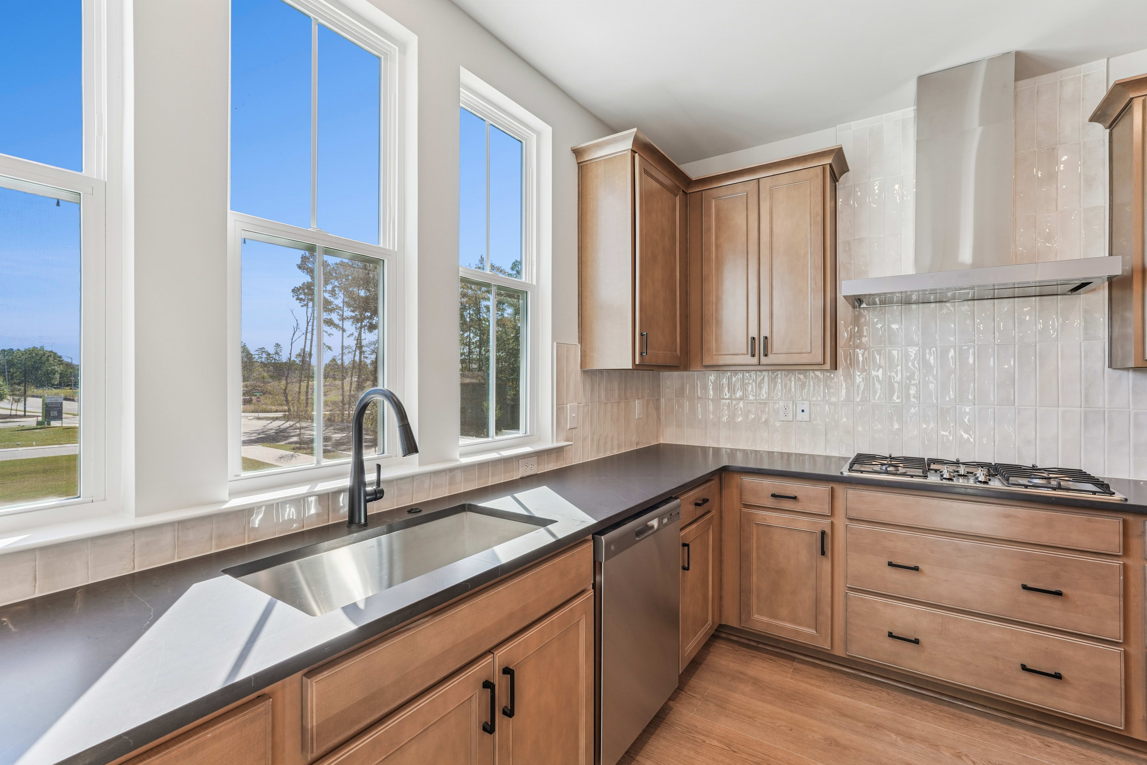 A modern, well-lit kitchen with wooden cabinets, a stainless steel sink, and large windows overlooking a scenic outdoor landscape.