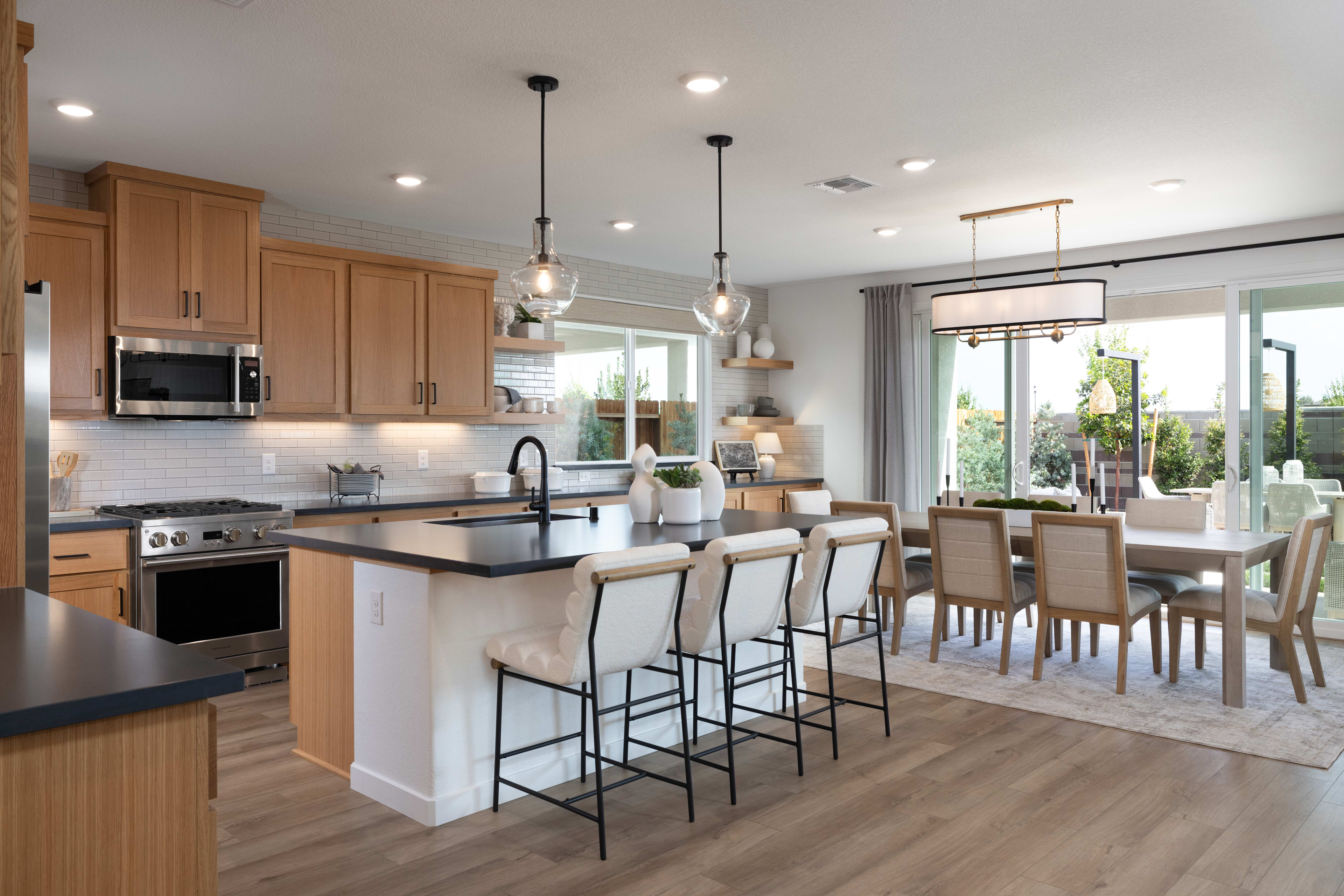 A modern, open-concept kitchen and dining area with light wood cabinets, black countertops, and pendant lighting over a central island and dining table.