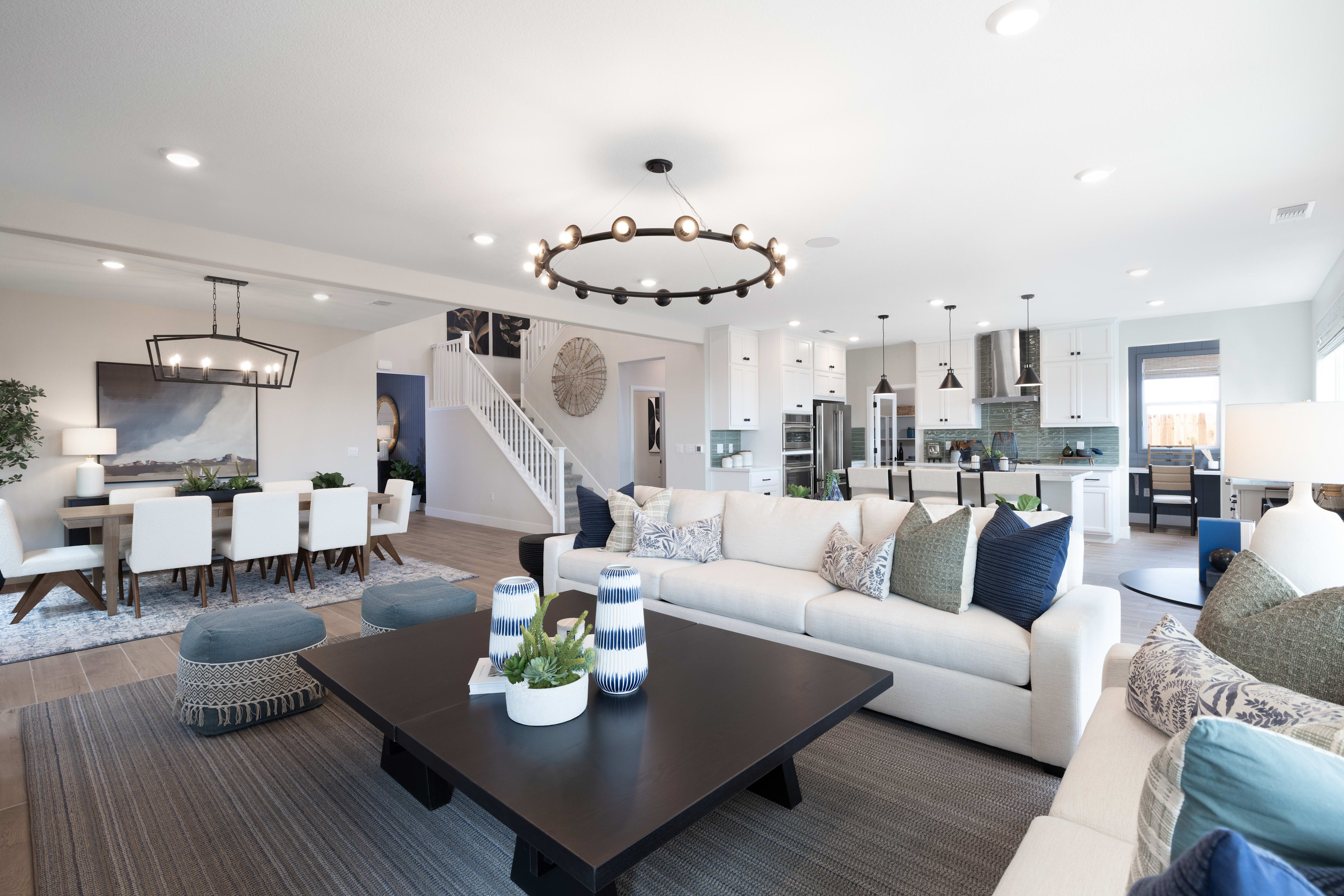 A spacious and well-designed living room with a large sofa, coffee table, and a chandelier overhead, surrounded by a dining area and an open-concept kitchen in the background.