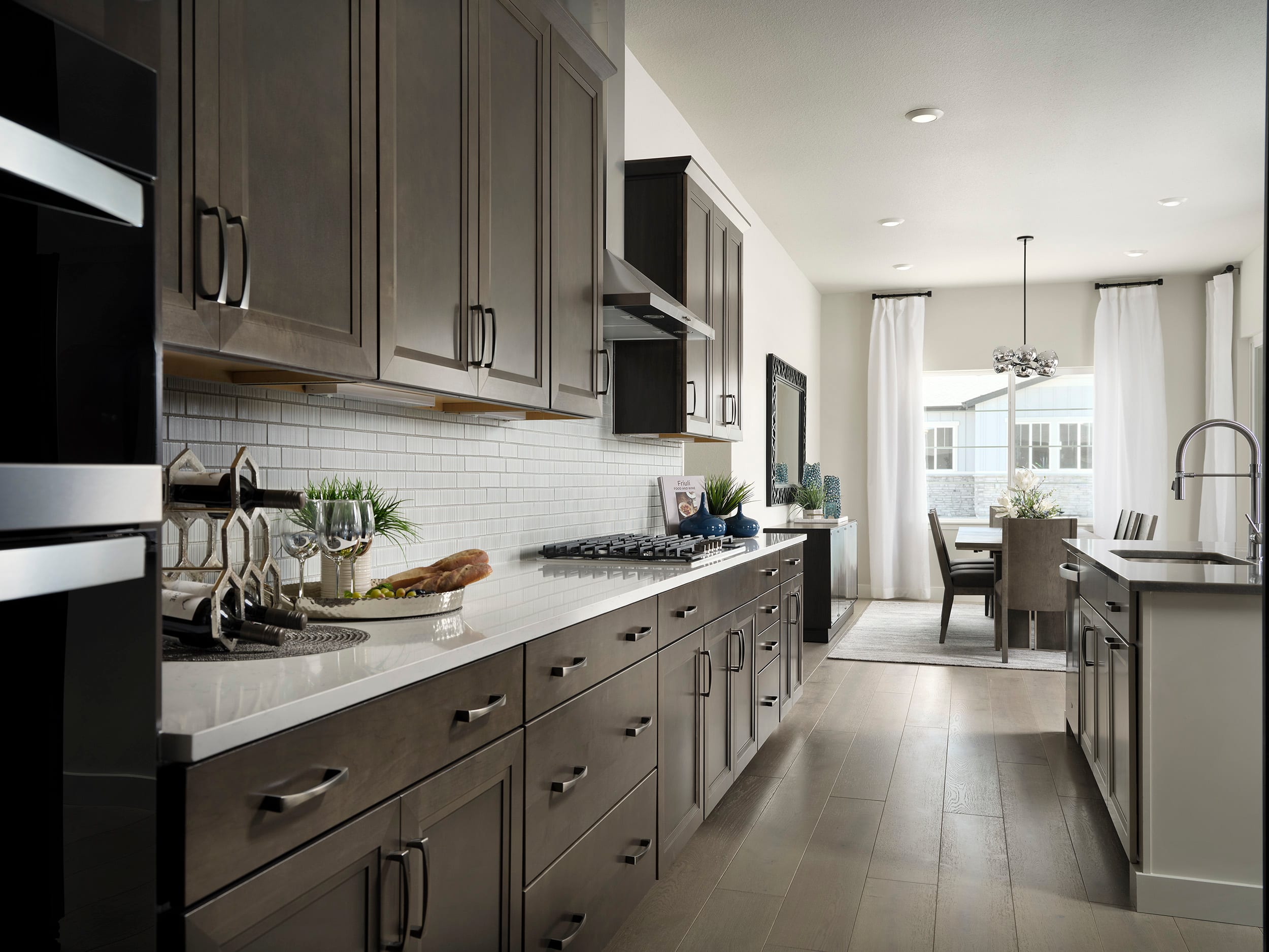 A modern, sleek kitchen with dark gray cabinets, white countertops, and stainless steel appliances, leading into a dining area with a chandelier and large windows.