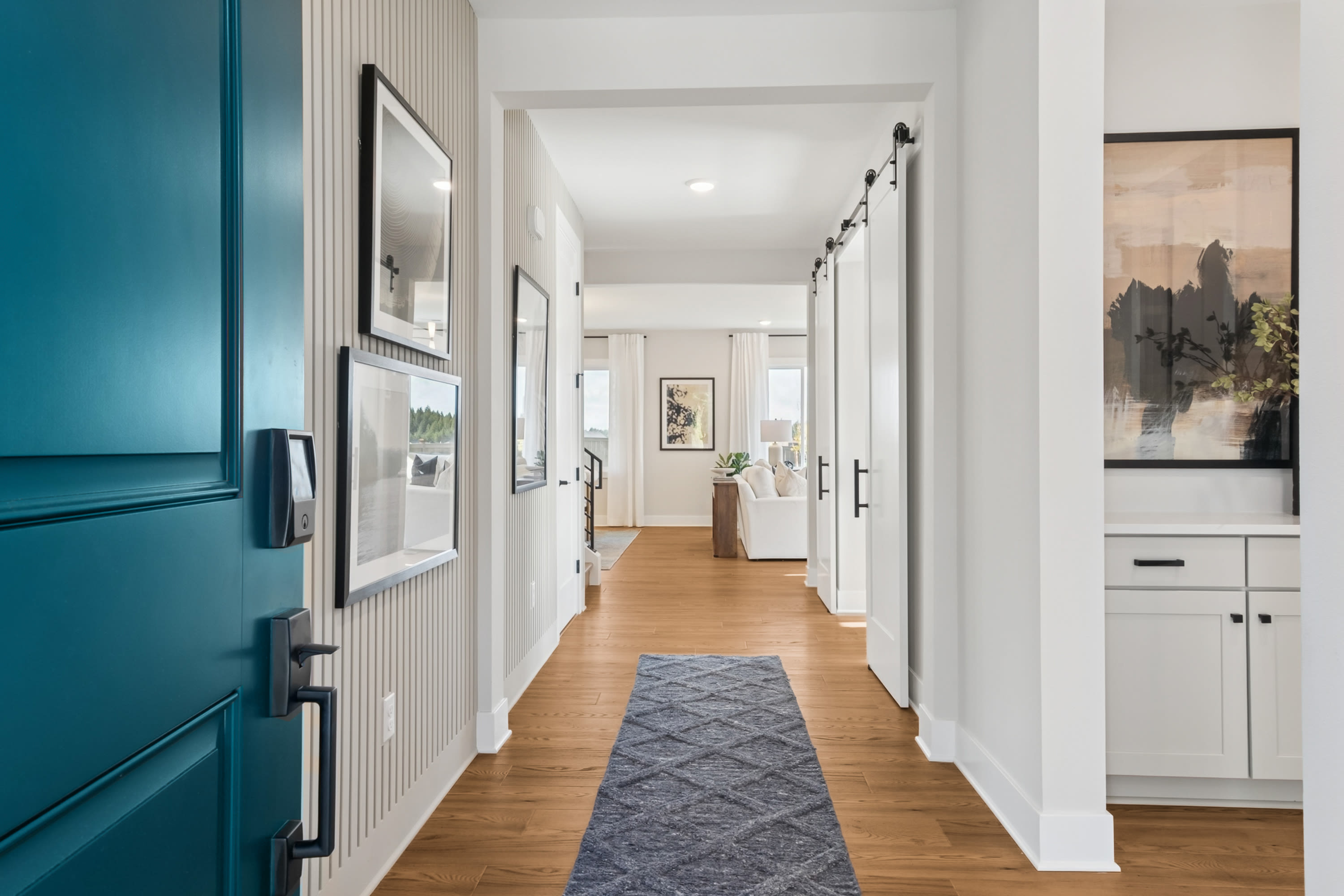 A bright and airy hallway with a teal-colored door, wooden floors, and artwork adorning the walls, leading to a kitchen area in the background.