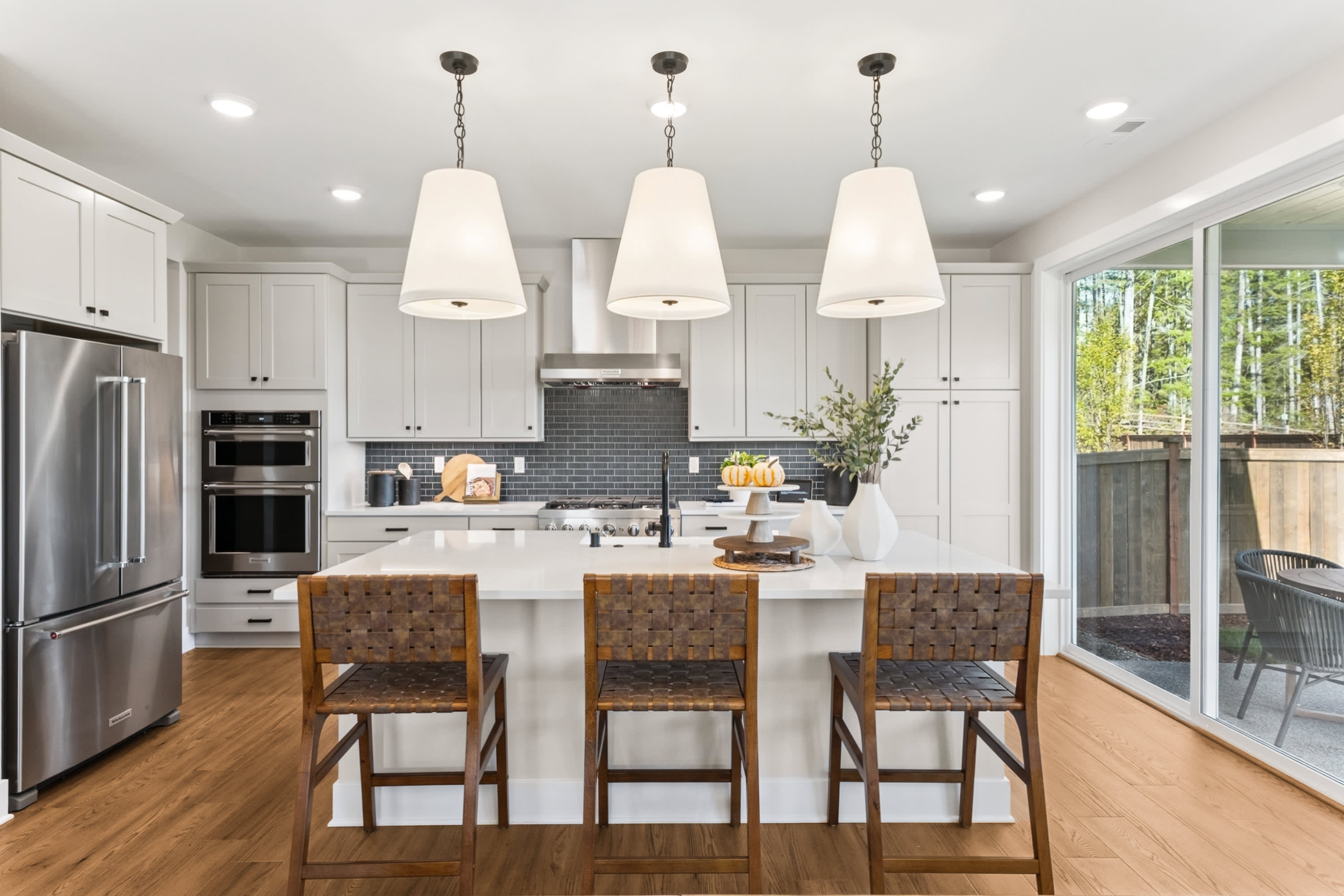 A modern, well-lit kitchen with white cabinets, a central island, and three pendant lights hanging above it. The kitchen has a hardwood floor and large windows overlooking a wooded area outside.