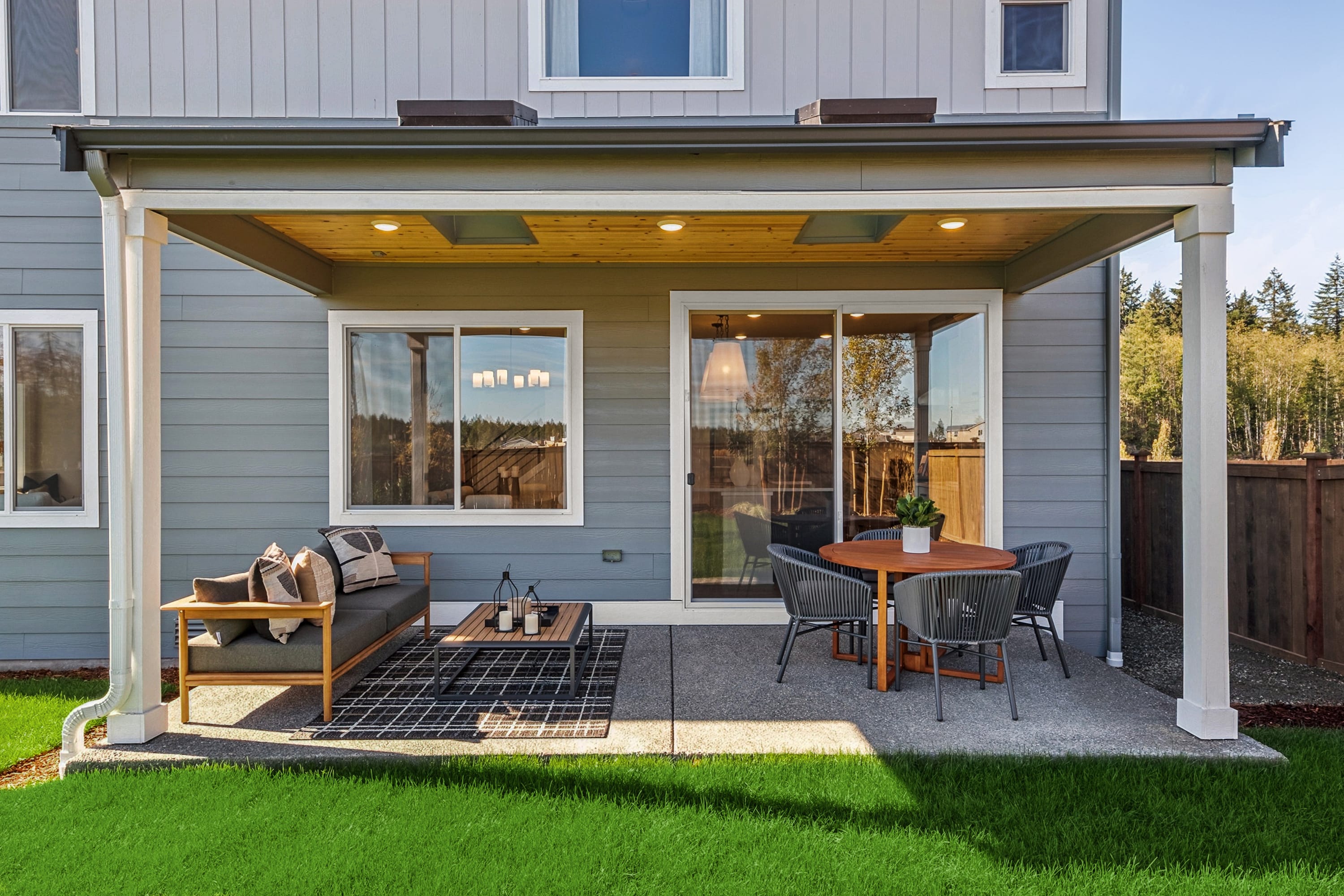 A cozy outdoor patio with a covered seating area, surrounded by a well-manicured lawn and a modern, gray-sided house in the background.