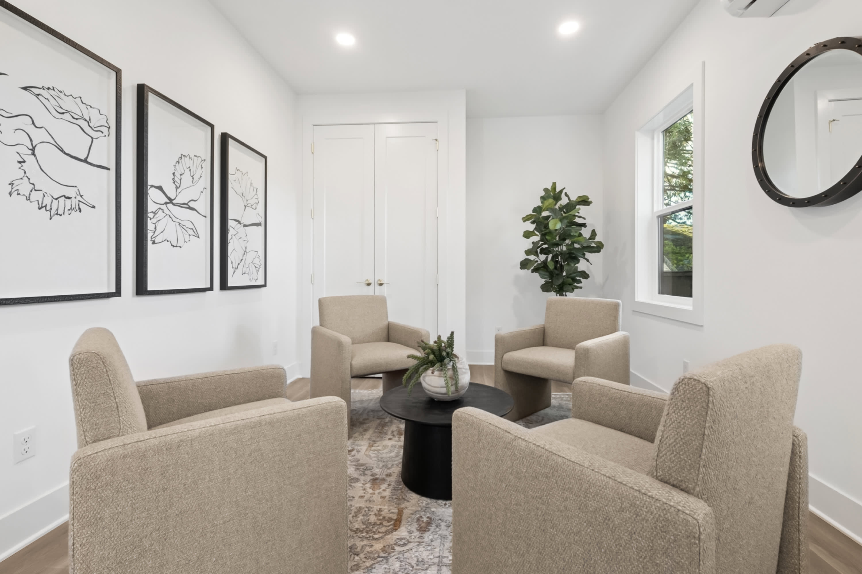 A cozy and modern living room with beige armchairs, a round coffee table, and framed artwork on the walls, complemented by a large window providing natural light.