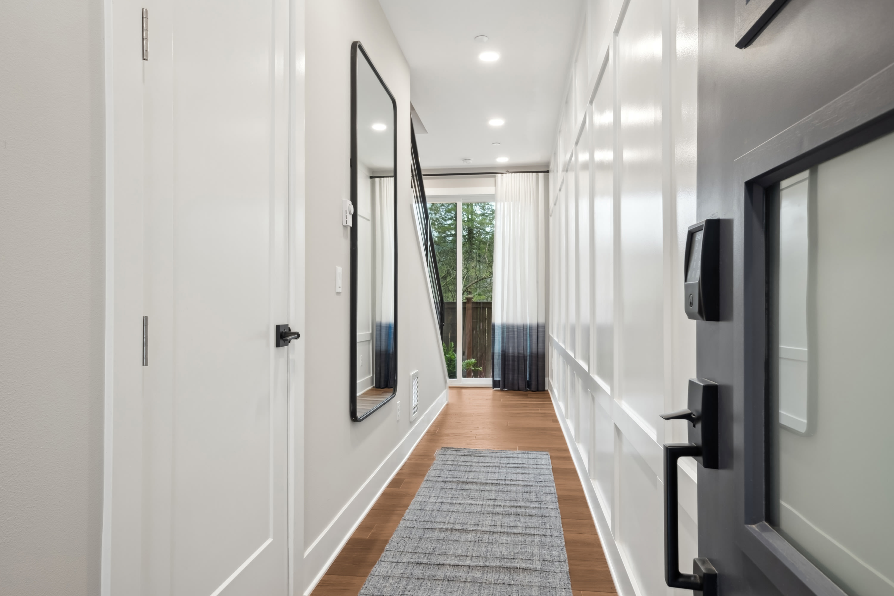 A bright, minimalist hallway with white walls, a wooden floor, and a large mirror hanging on the wall, leading to a window that provides natural light.