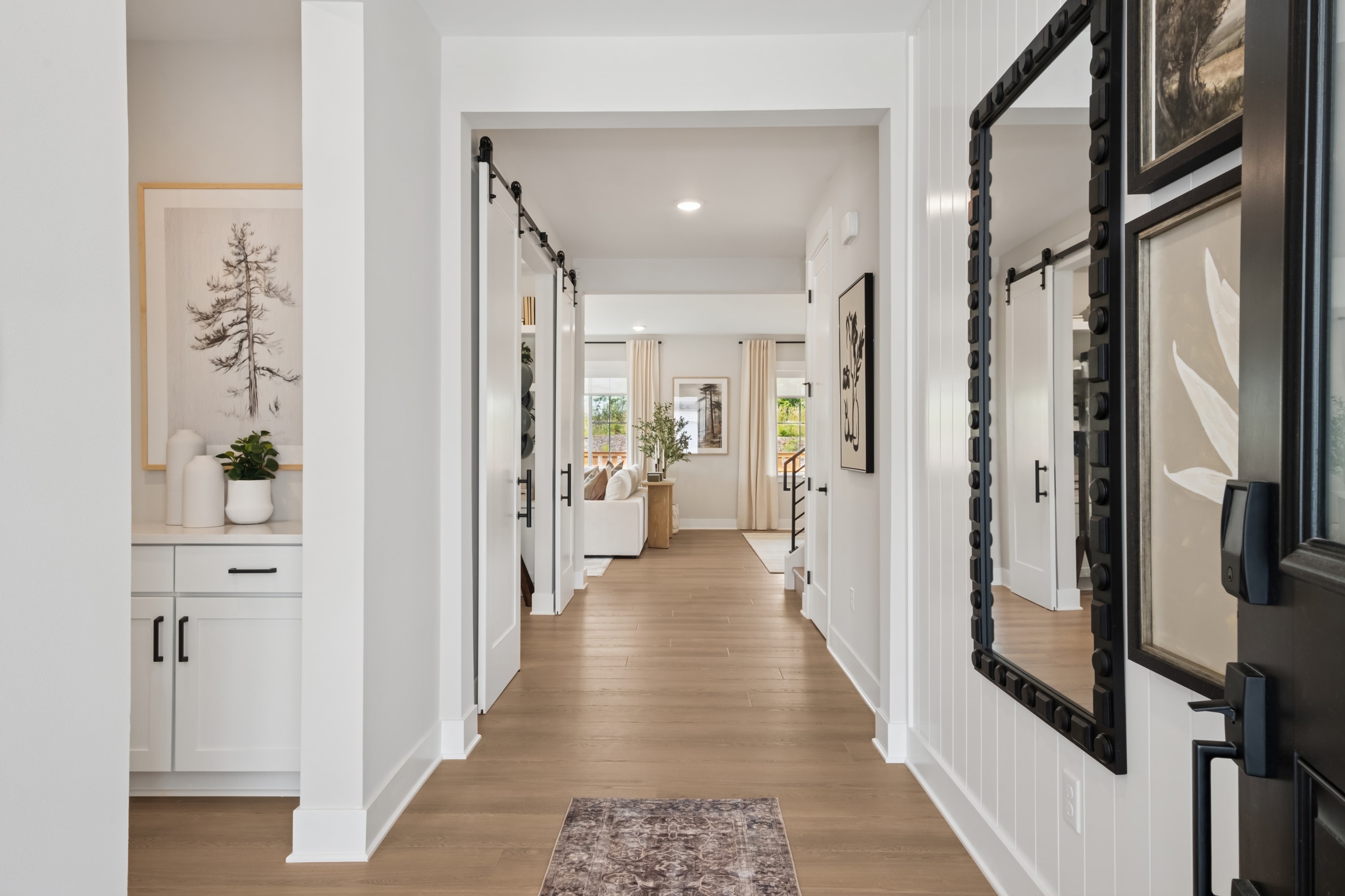 A bright and airy hallway with white walls, wooden floors, and a decorative rug leading the way to other rooms, with framed artwork adorning the walls.