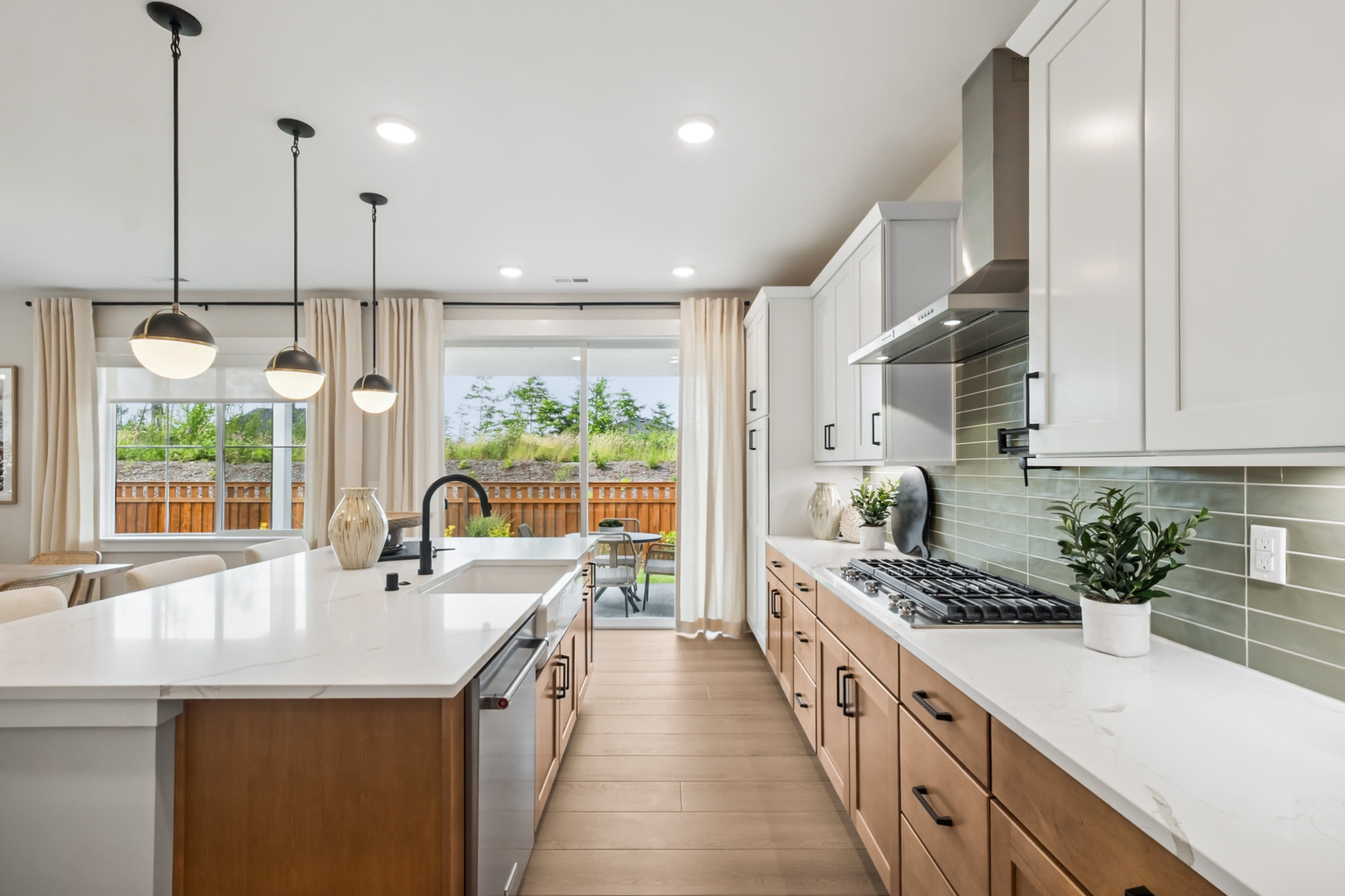 A modern, open-concept kitchen with white countertops, wooden cabinets, and pendant lighting, overlooking a backyard through large windows.