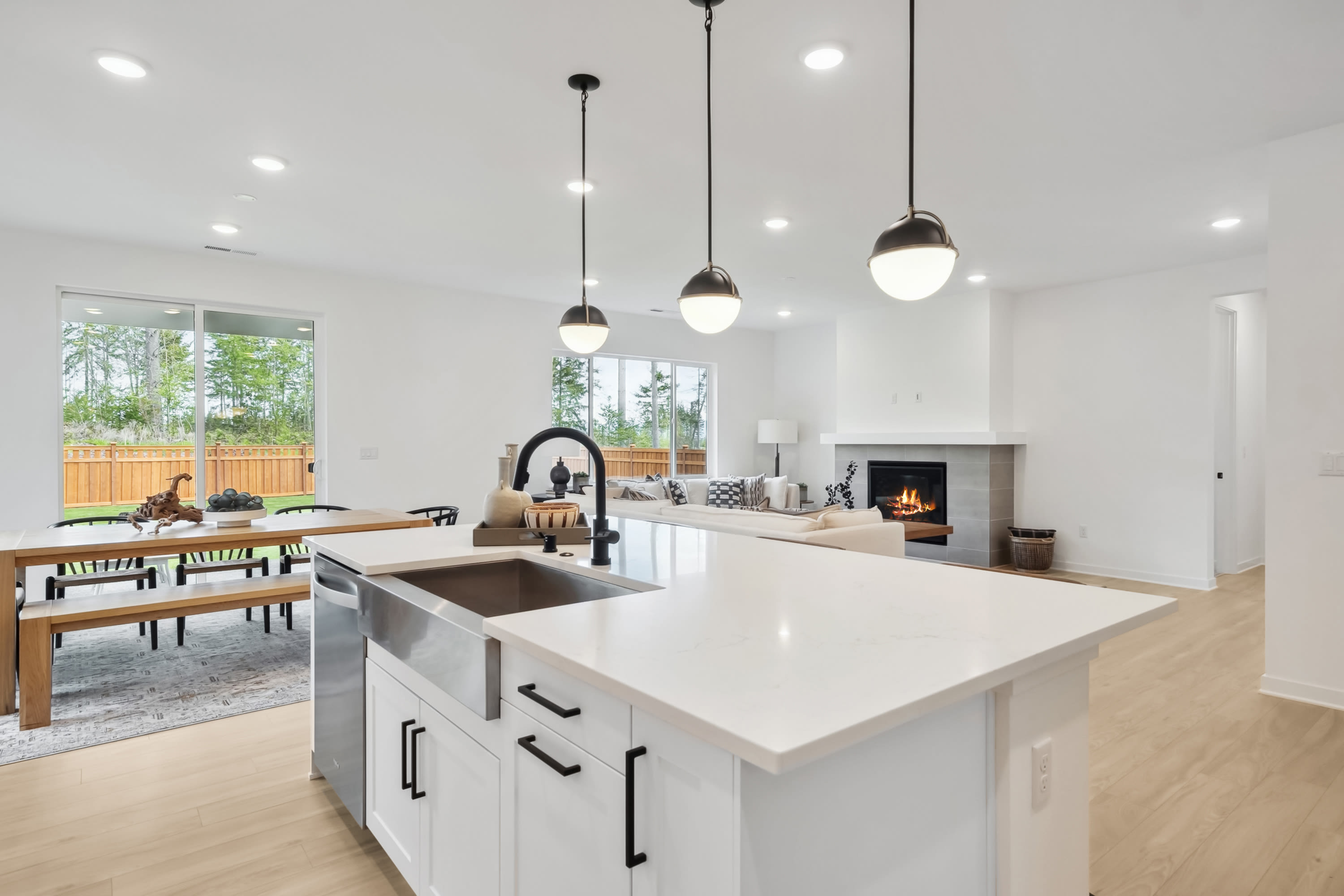 A modern, open-concept kitchen with a large island, pendant lighting, and a fireplace visible in the background, surrounded by a wooden fence and greenery outside.