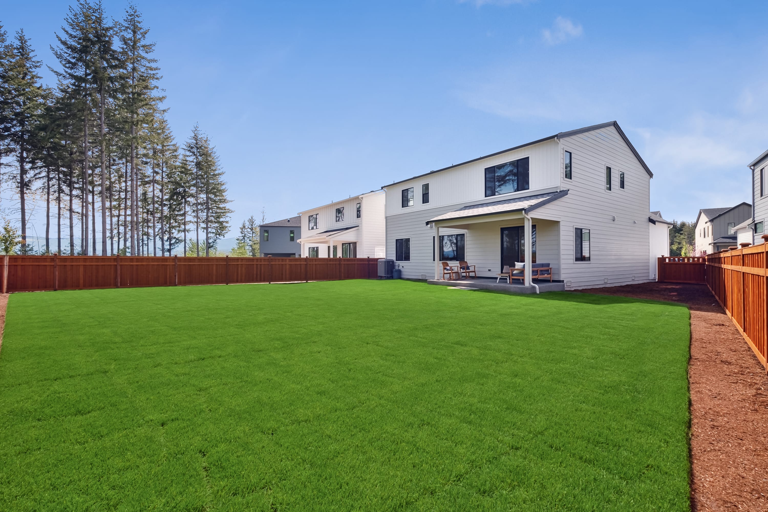 A well-manicured green lawn in the foreground leads to a modern two-story residential building with a wooden fence and tall pine trees in the background.