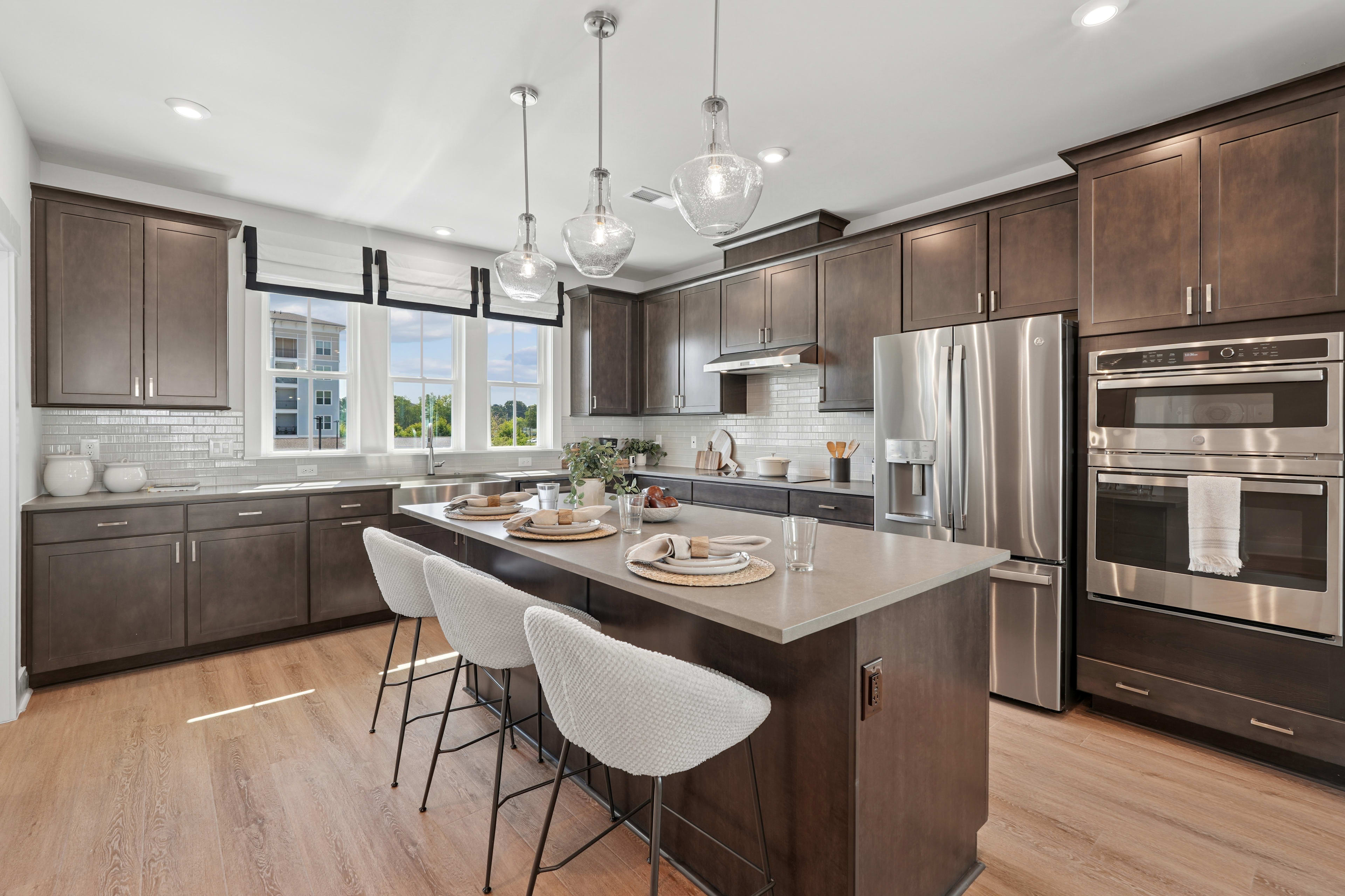 A modern and spacious kitchen with dark wood cabinets, stainless steel appliances, a large island with bar stools, and pendant lighting fixtures.