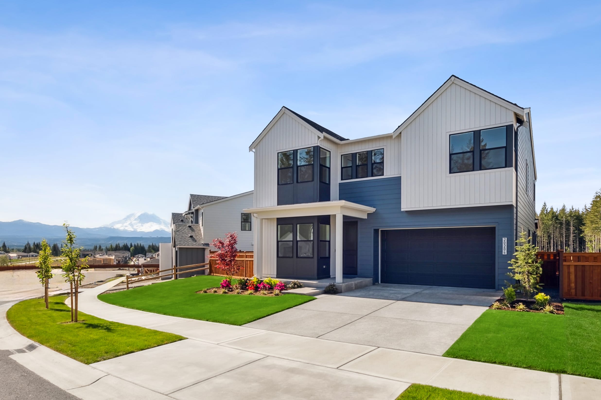 A modern two-story house with a garage, surrounded by a well-manicured lawn and landscaping, set against a backdrop of mountains and a clear blue sky.