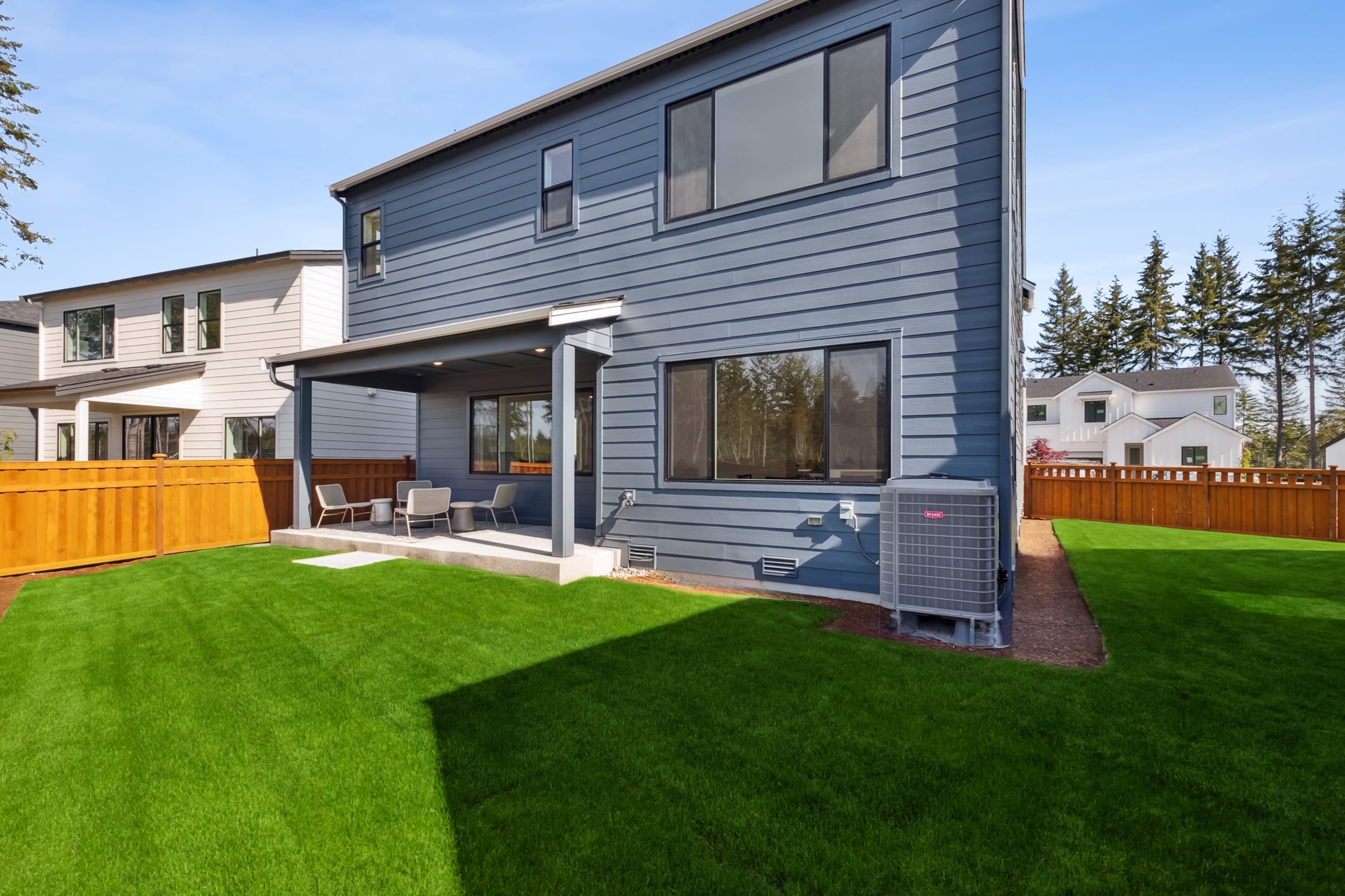 A modern two-story house with gray siding, a wooden deck, and a well-manicured lawn in the foreground, surrounded by pine trees in the background.
