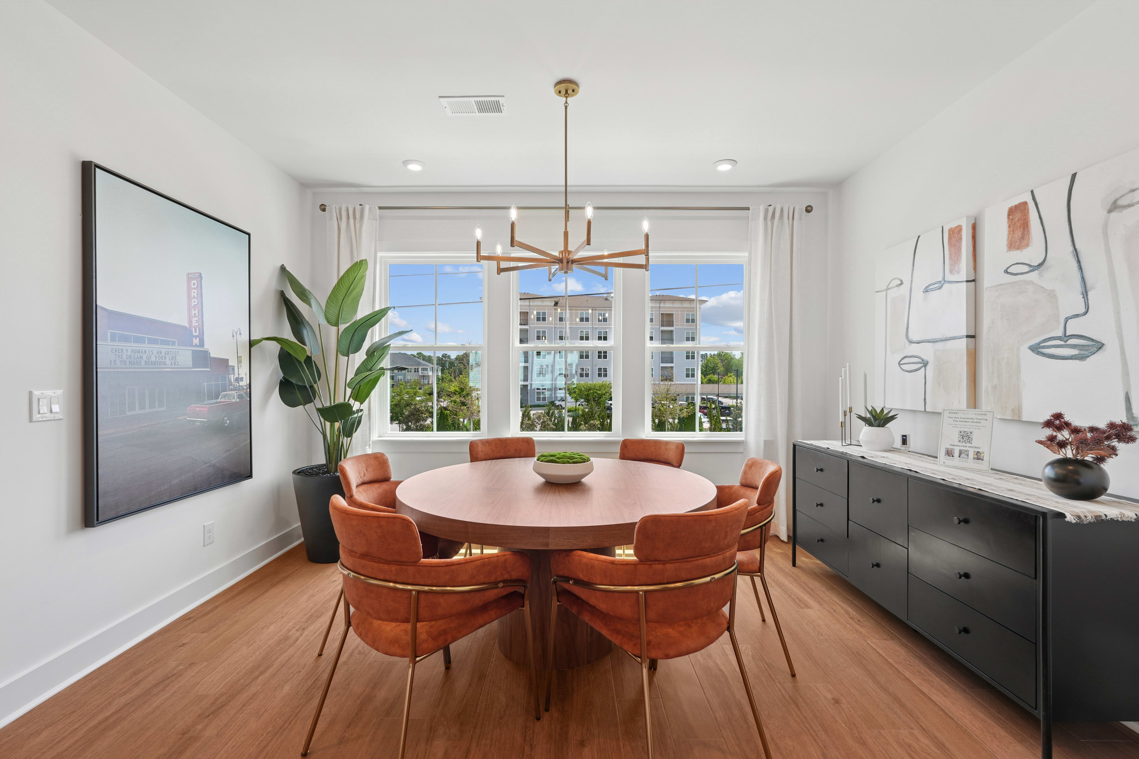 A modern and stylish dining area with a round wooden table surrounded by plush orange chairs, set against a backdrop of large windows overlooking an urban landscape.