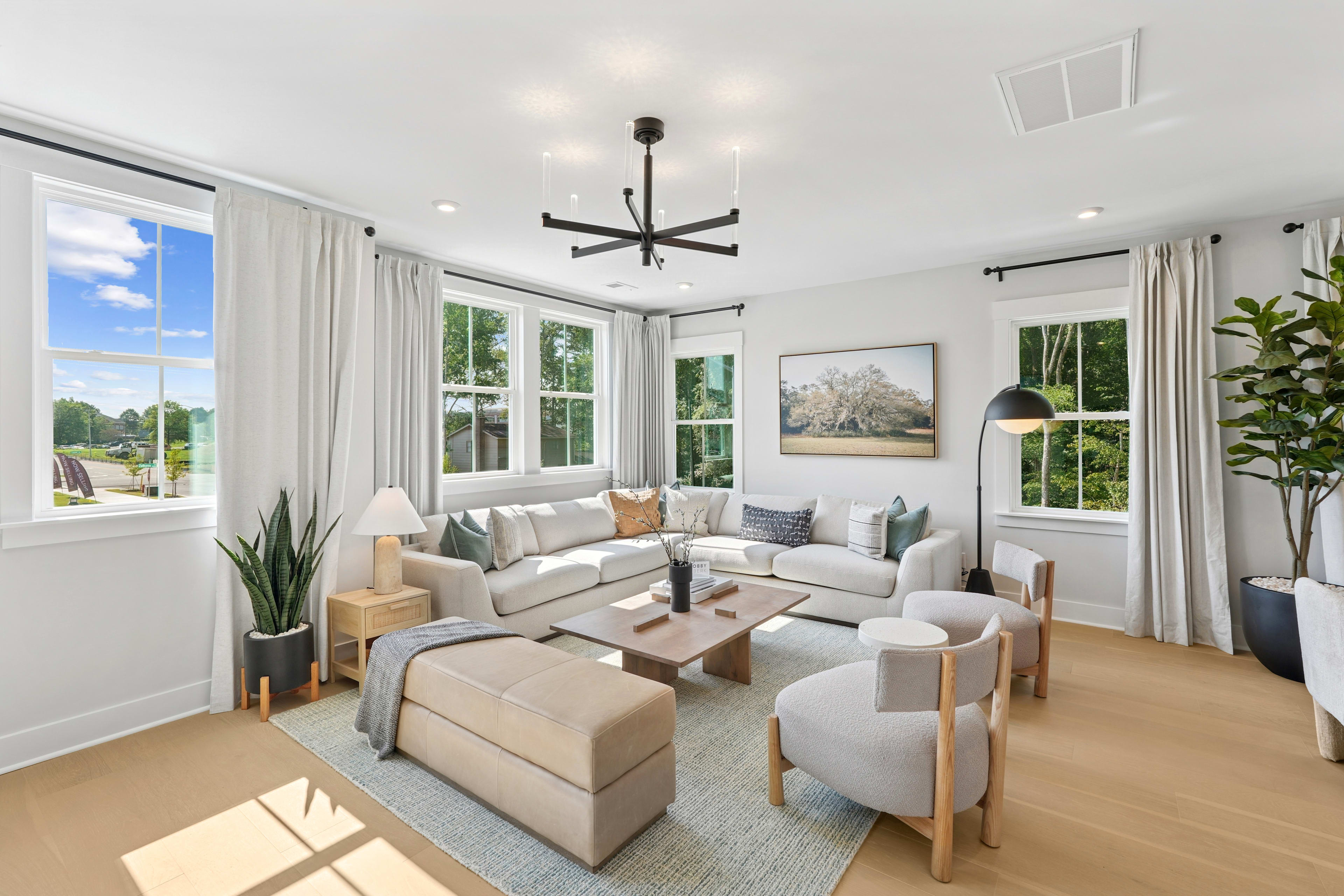 A spacious and well-lit living room with a comfortable sectional sofa, armchairs, and a wooden coffee table, surrounded by large windows that offer a scenic view of the outdoor greenery.
