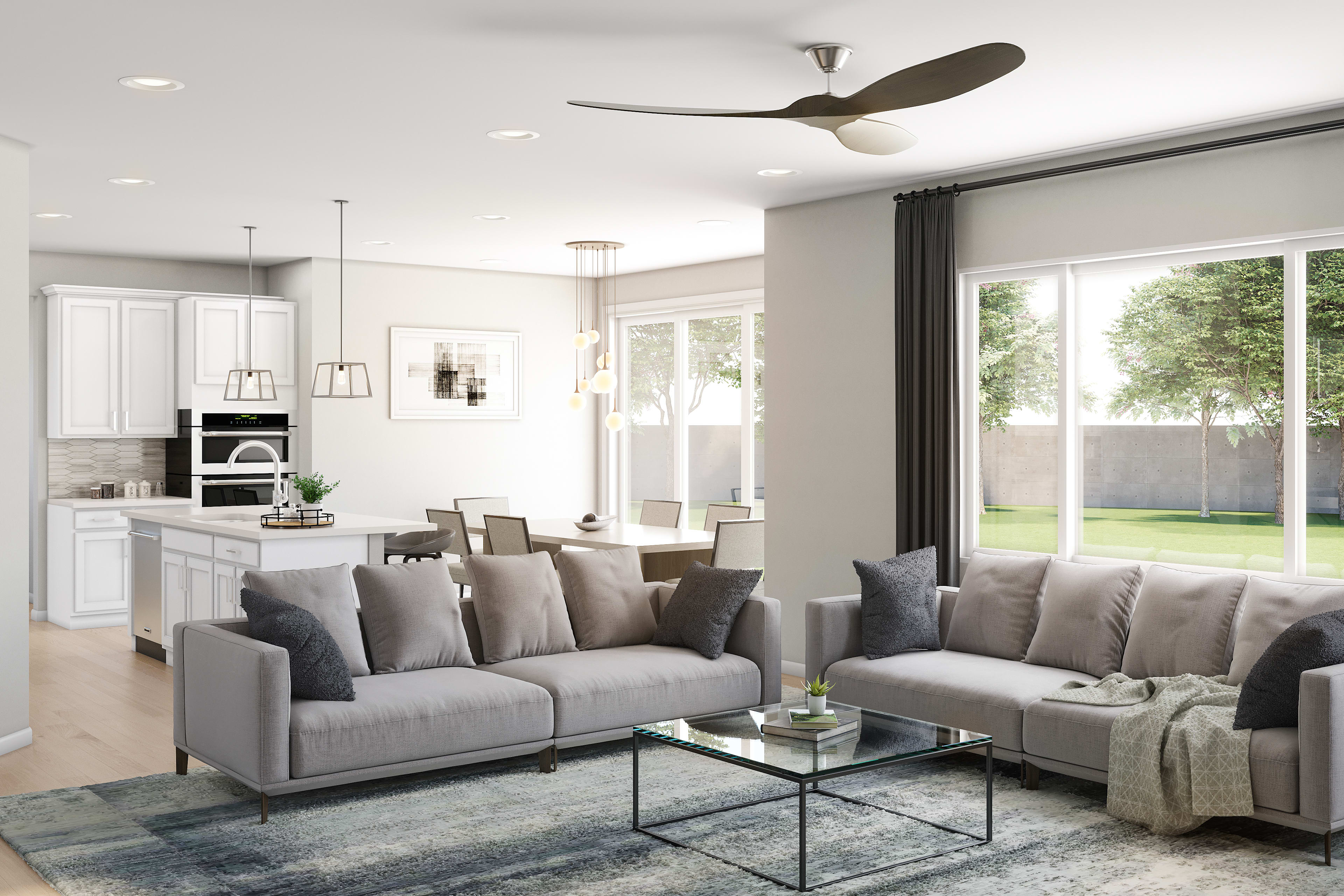 A modern and spacious living room with a gray sectional sofa, a glass coffee table, and a ceiling fan, surrounded by large windows overlooking a backyard.
