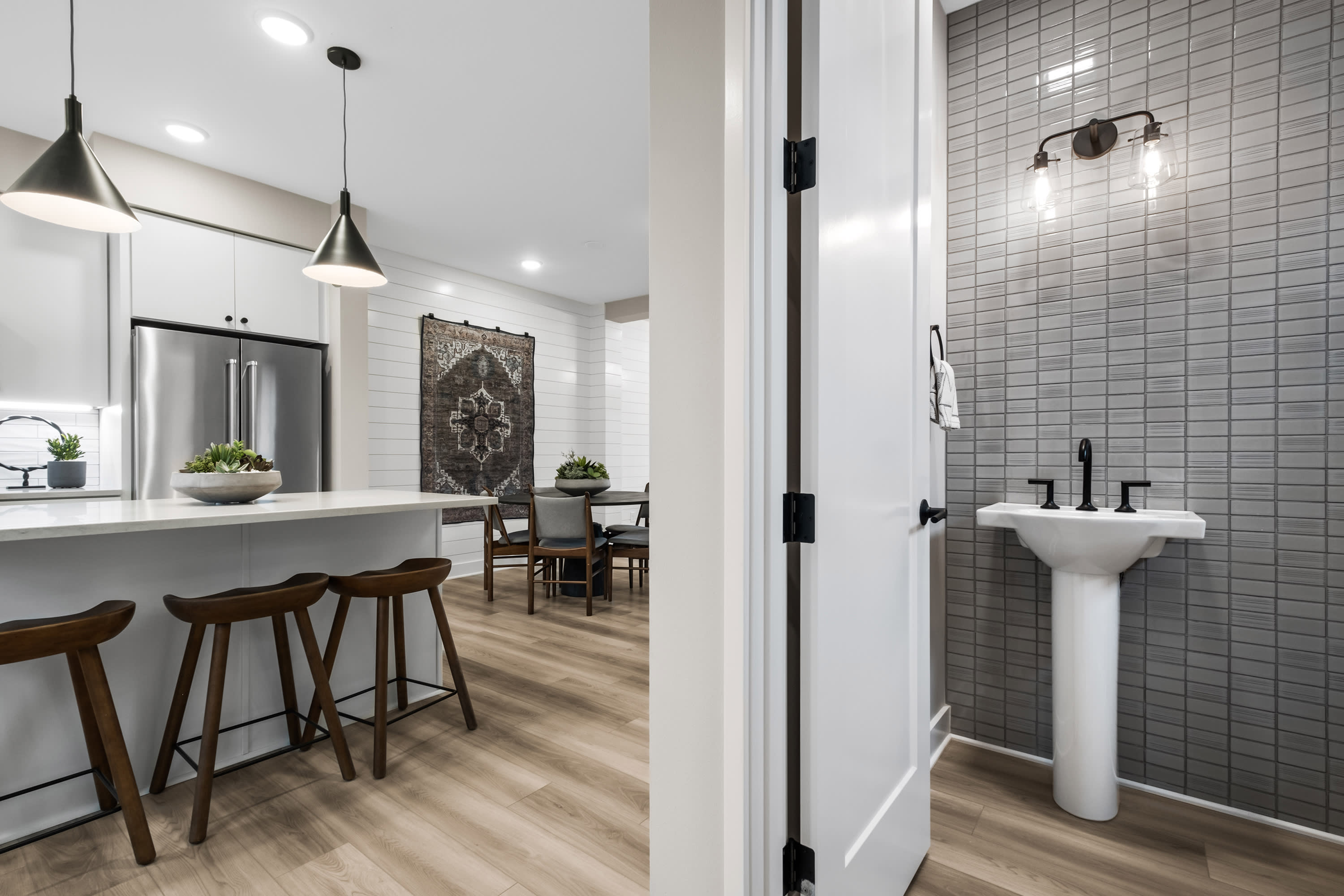 A modern, open-concept kitchen and dining area with a sleek, tiled bathroom visible in the background, featuring a pedestal sink and dark fixtures.