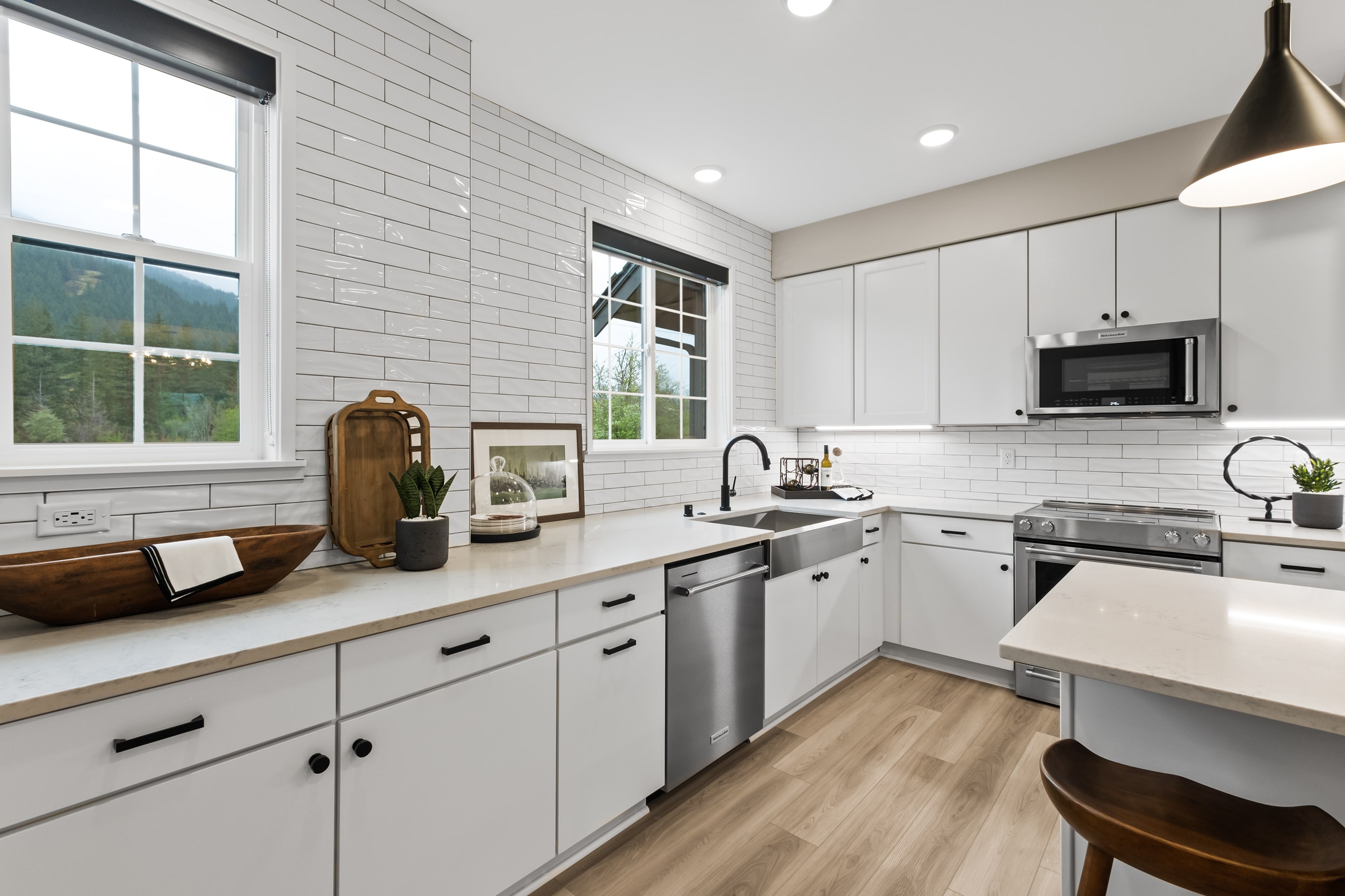 A modern, well-lit kitchen with white cabinets, stainless steel appliances, and a wooden countertop, set against a backdrop of a brick wall and large windows overlooking a scenic outdoor view.