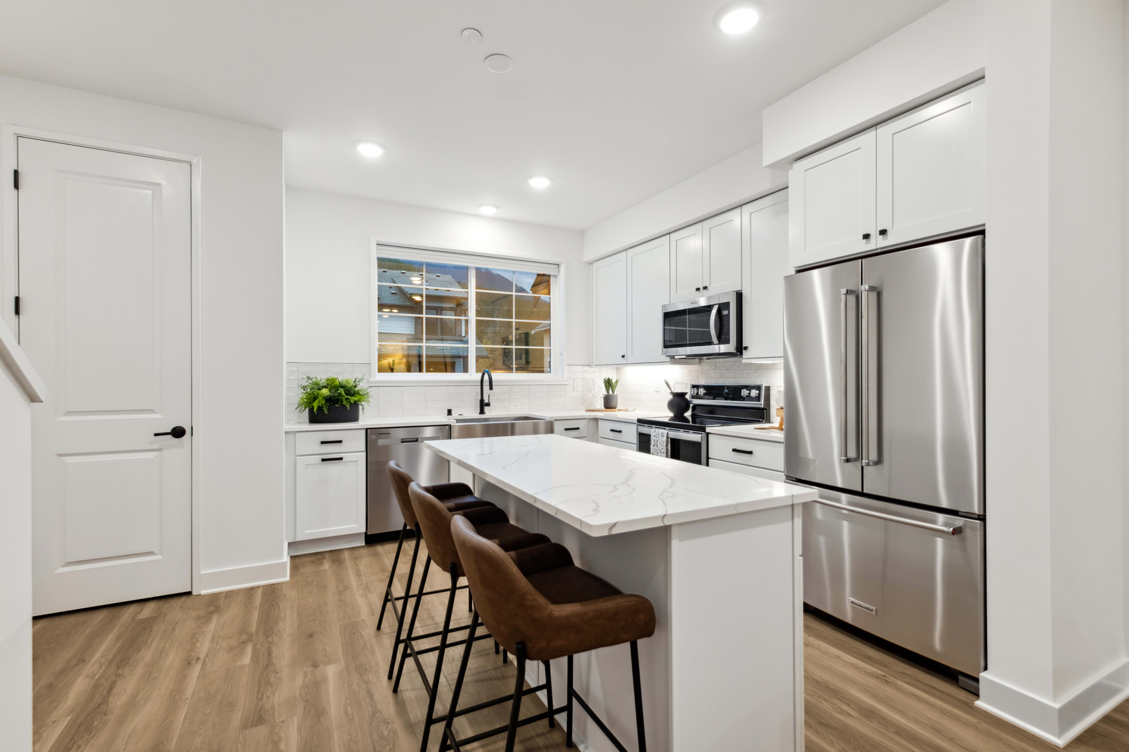 A modern, well-lit kitchen with white cabinets, stainless steel appliances, and a marble-topped island with bar stools in the foreground.