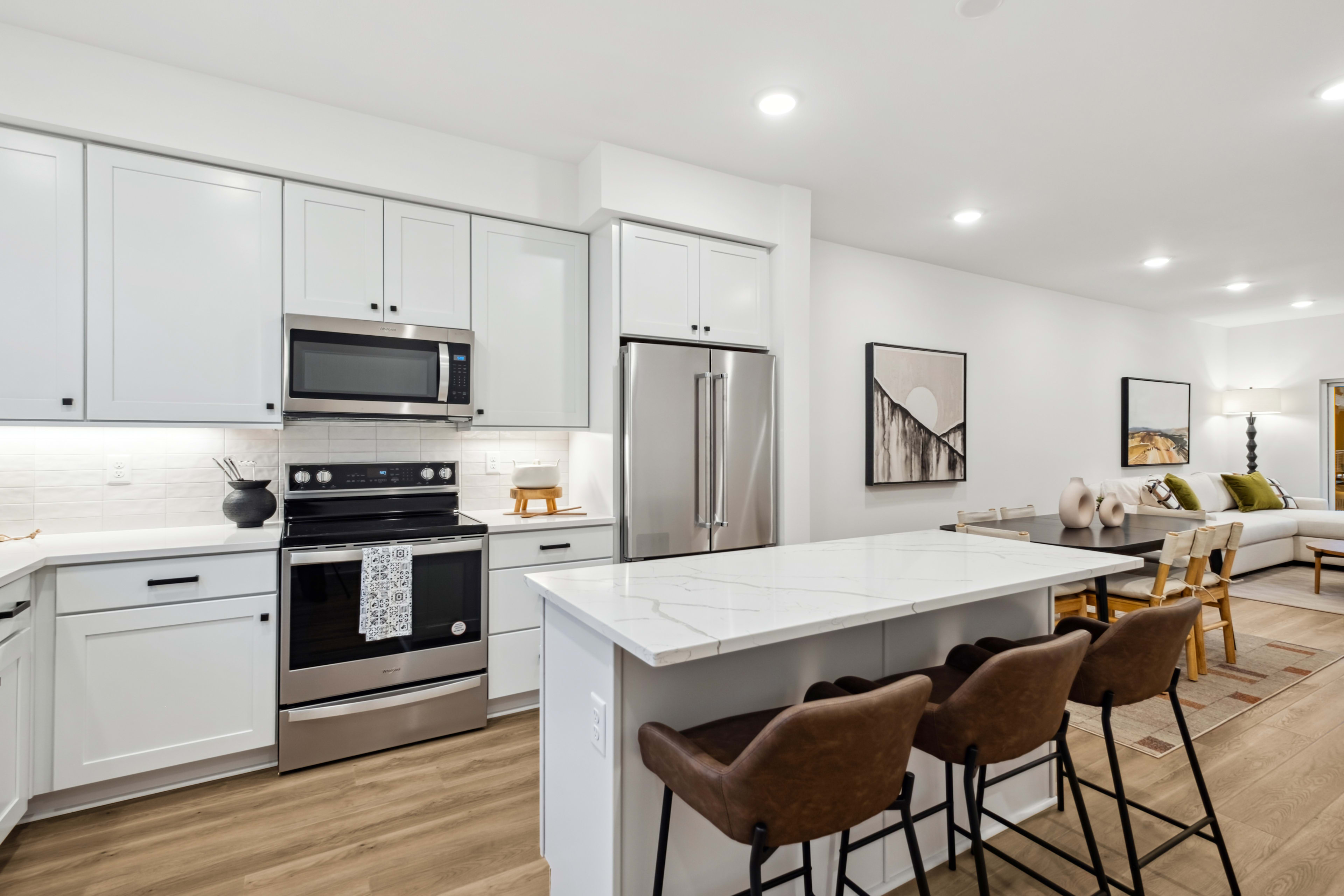 A modern, open-concept kitchen with white cabinets, stainless steel appliances, and a large central island with bar stools, set against a backdrop of a living room area with a couch.