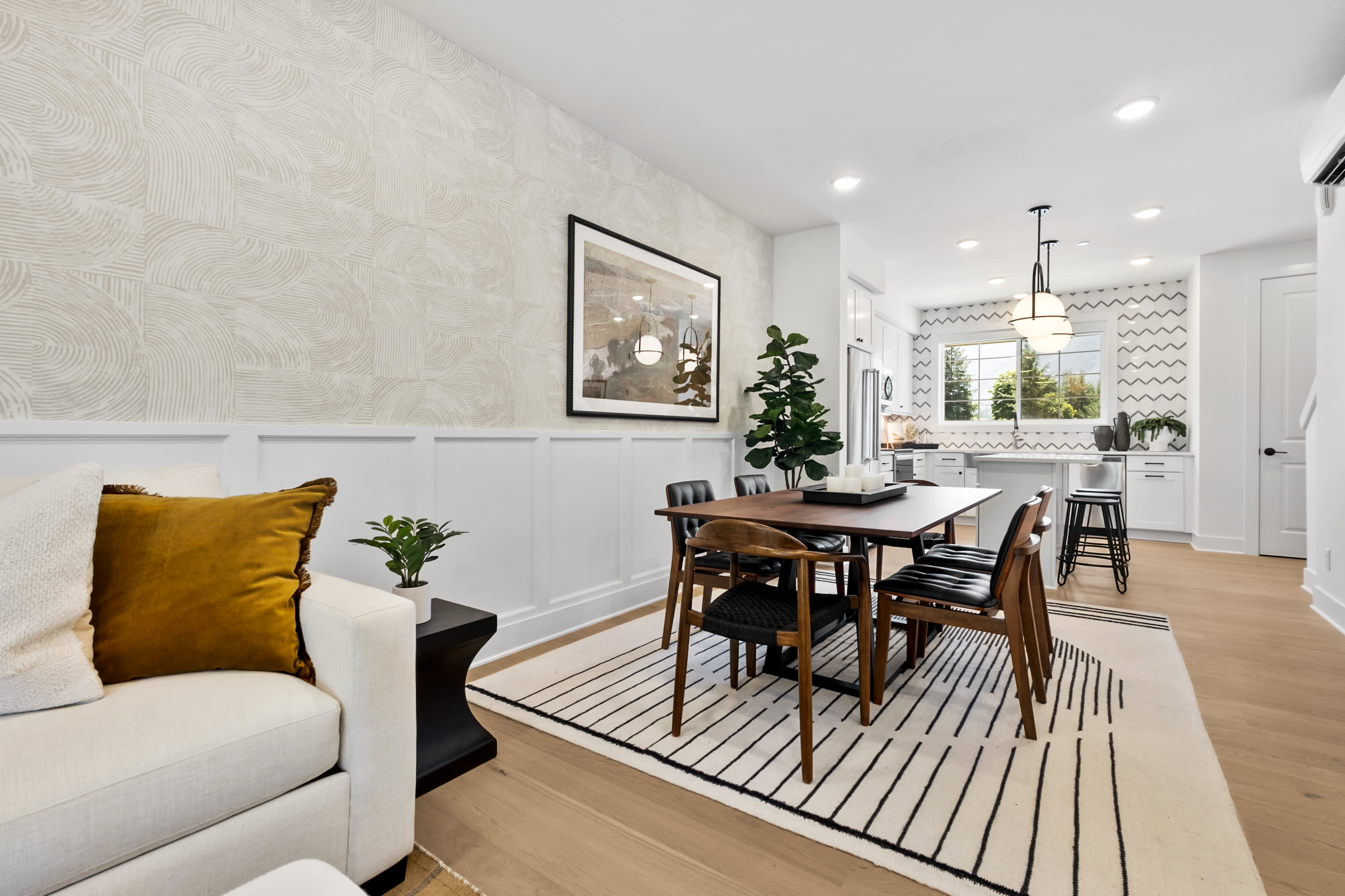 A modern and minimalist dining area with a white sofa, a wooden dining table and chairs, and various decorative elements such as a framed artwork and potted plants.