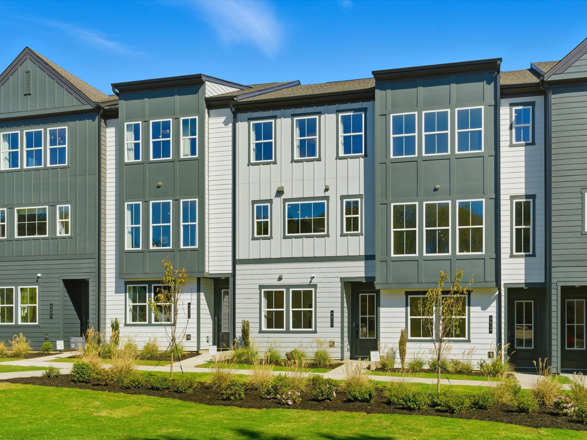 A row of modern, multi-story townhouses with green and gray siding, surrounded by a well-manicured lawn and landscaping in the foreground, set against a clear blue sky in the background.