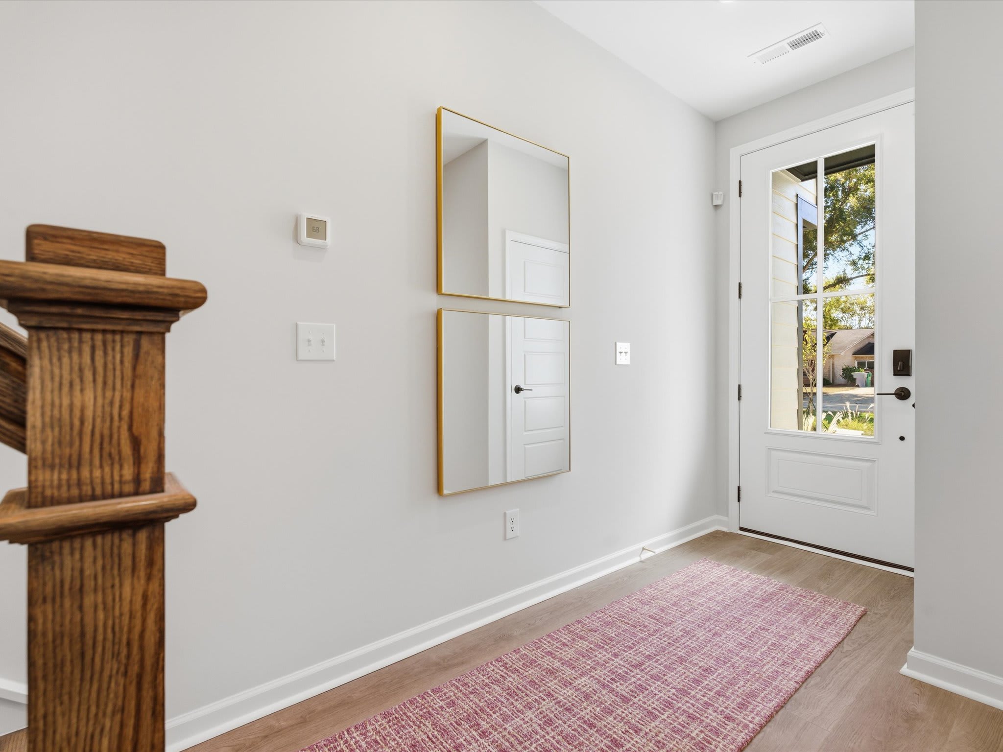A bright and airy entryway with a wooden staircase, two framed mirrors on the wall, and a pink patterned rug on the floor leading to a white door.